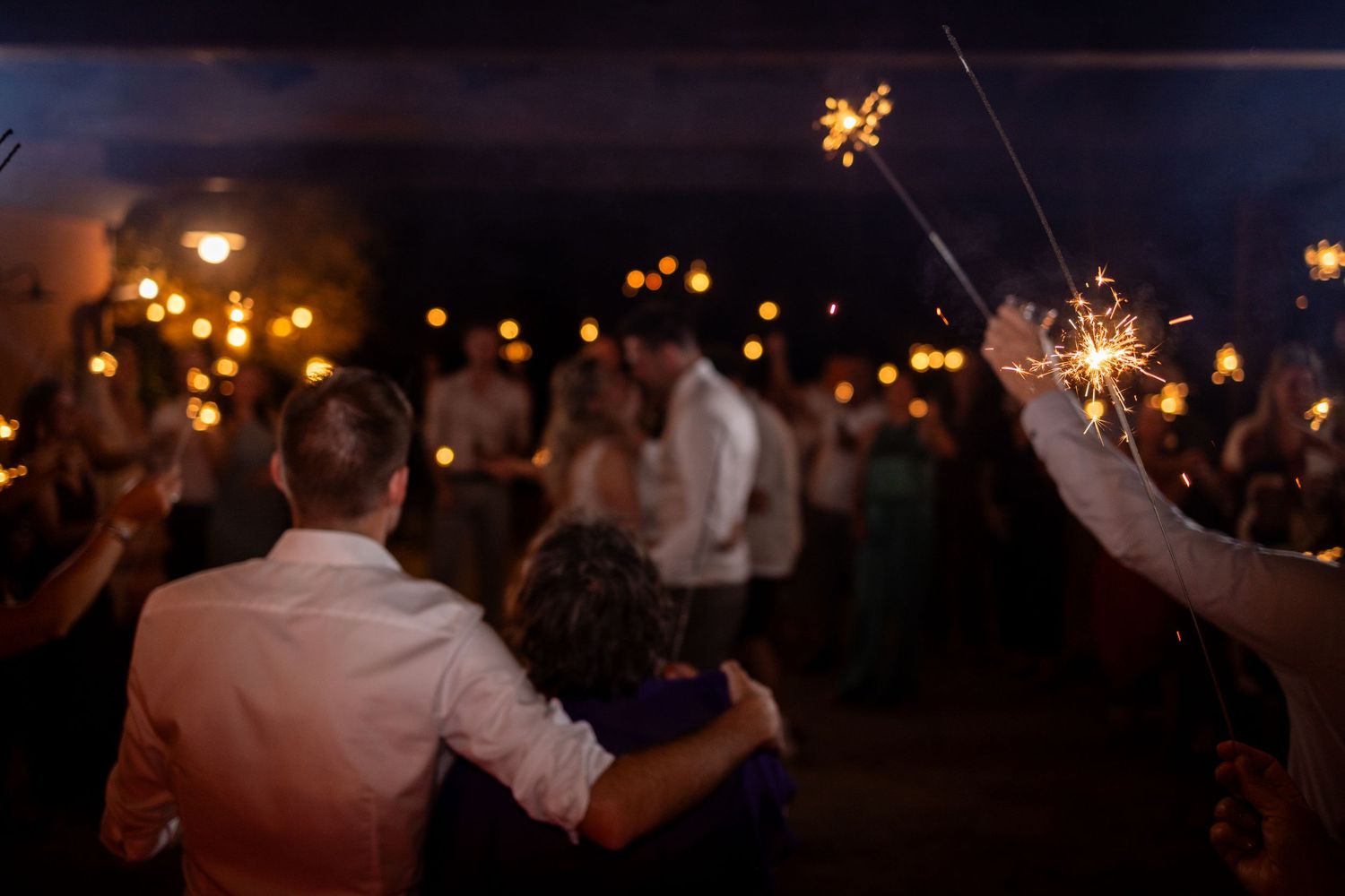Wedding guests celebrate with sparklers during an evening outdoor reception under glowing lights.