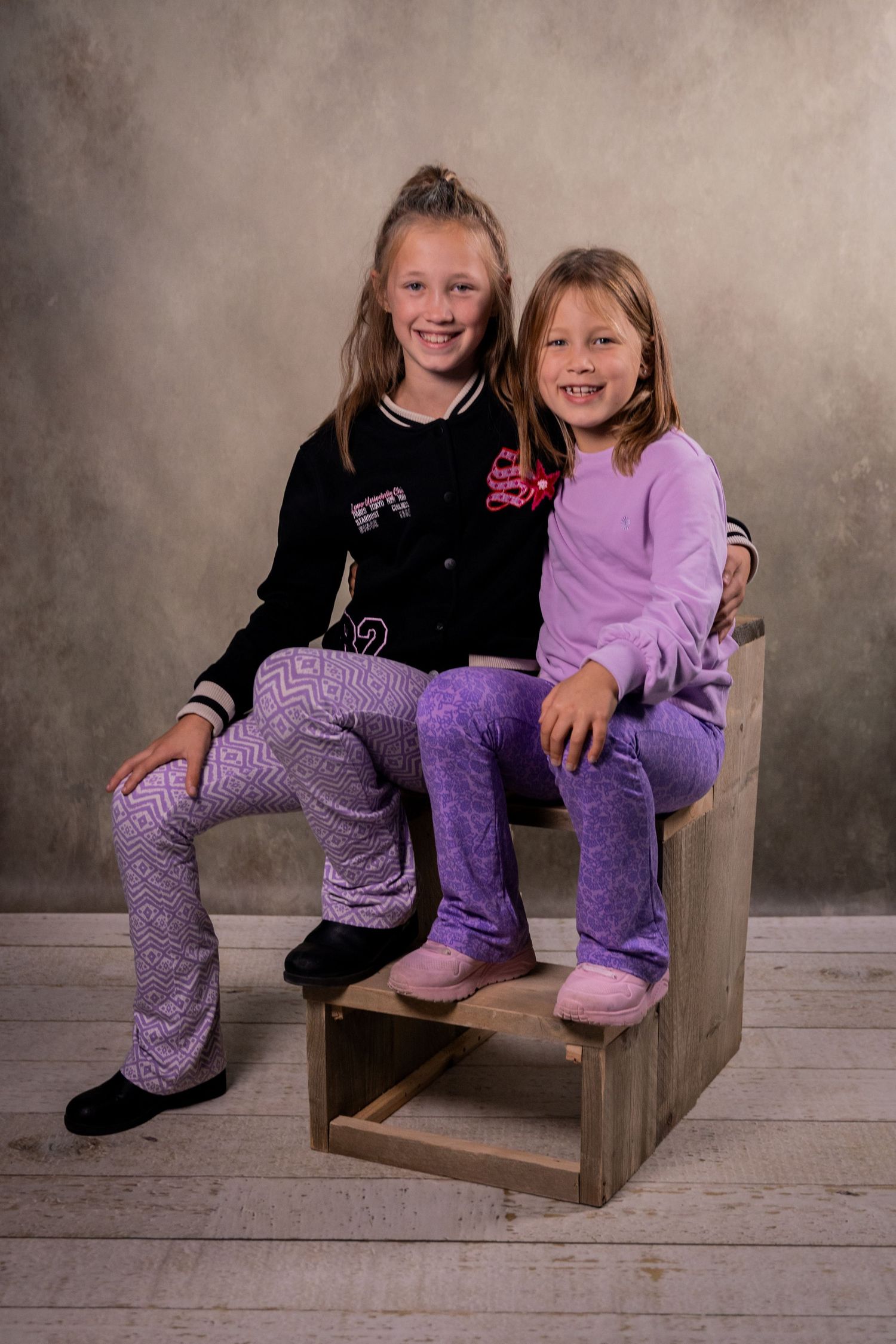 Two children in purple outfits sitting on wooden steps against a neutral backdrop.