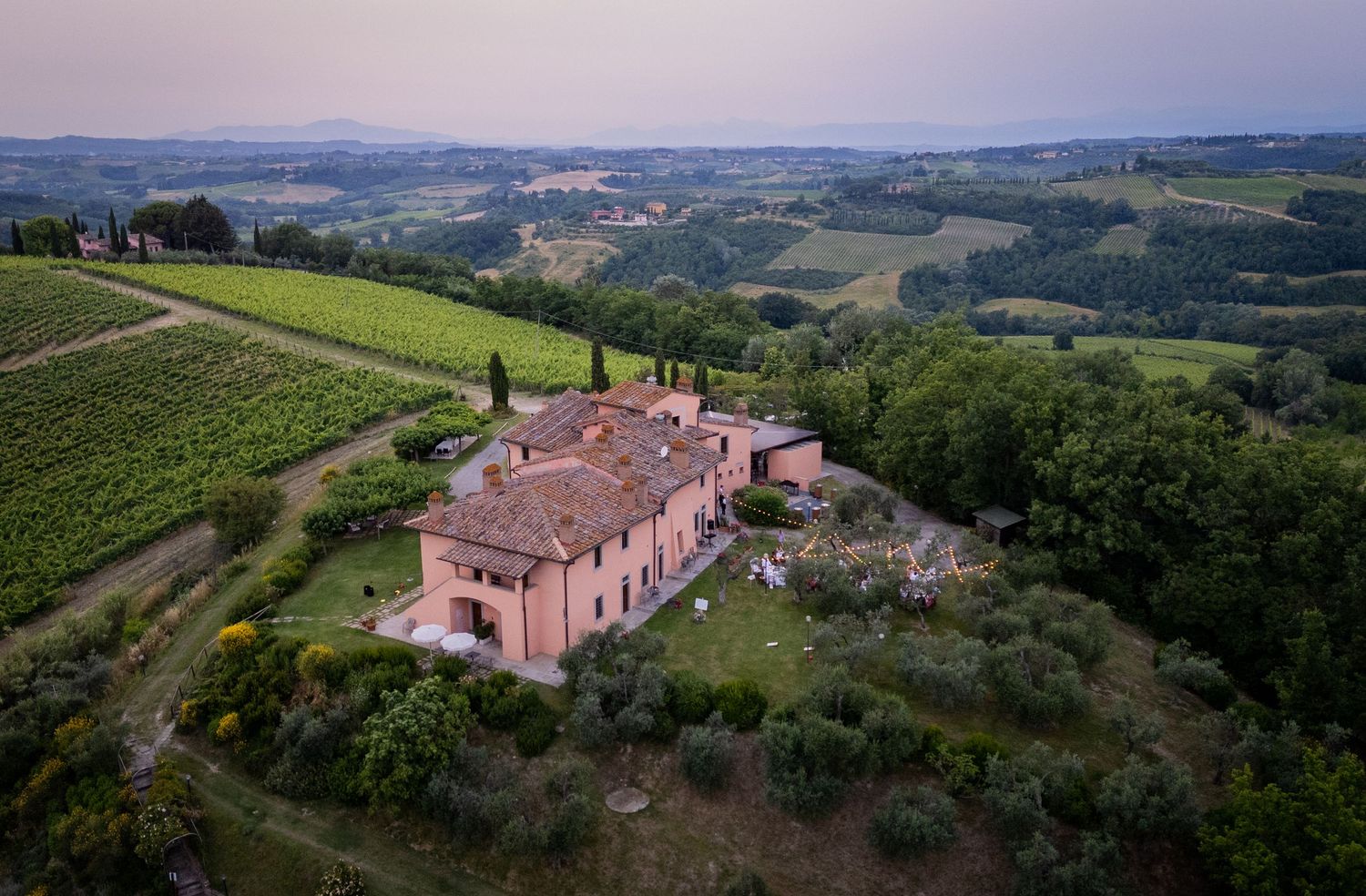 Aerial view of a rustic pink Tuscan villa perched on a hilltop surrounded by rolling green vineyards and countryside.