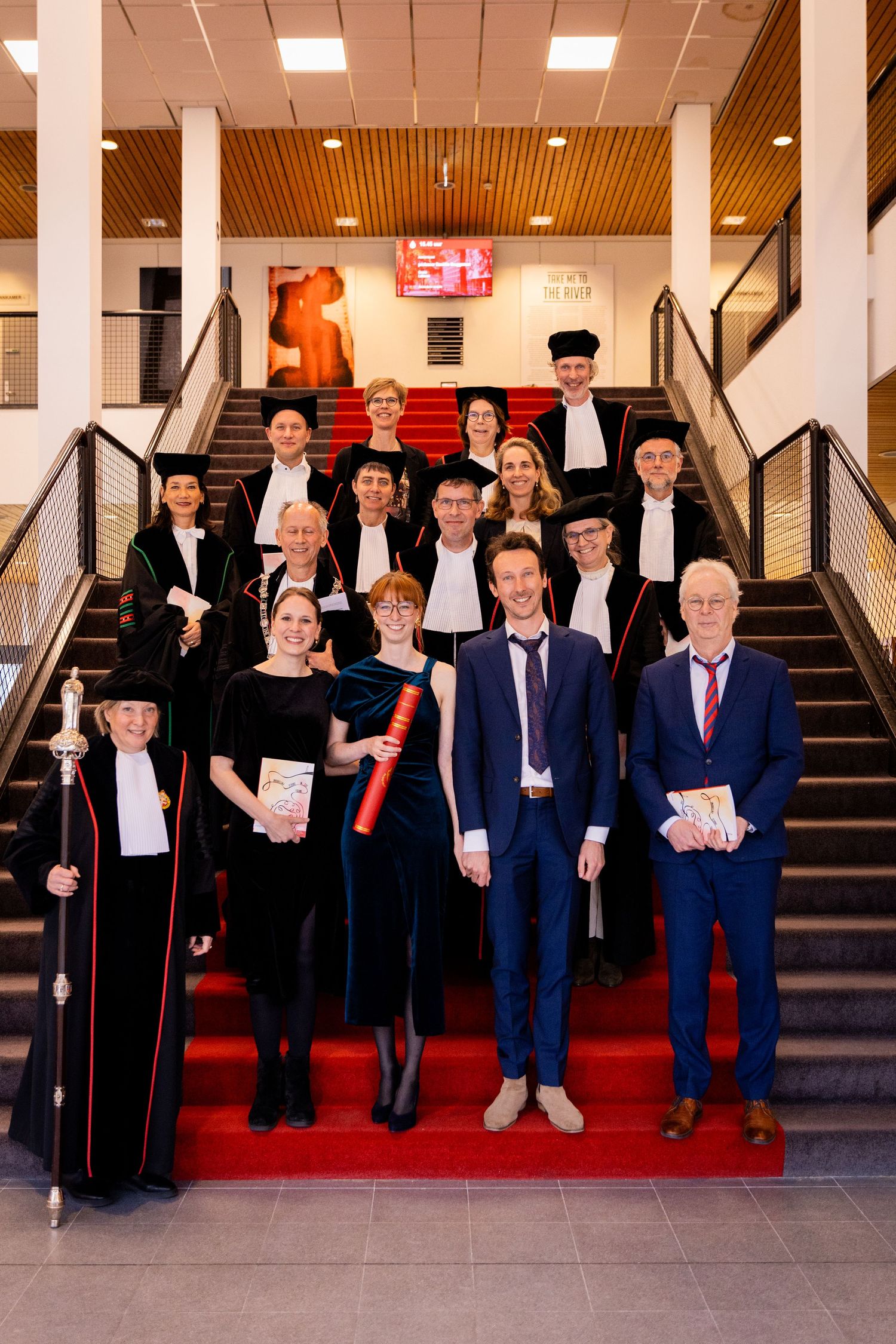 A formal group photo of graduates posed on red-carpeted stairs in an academic building.