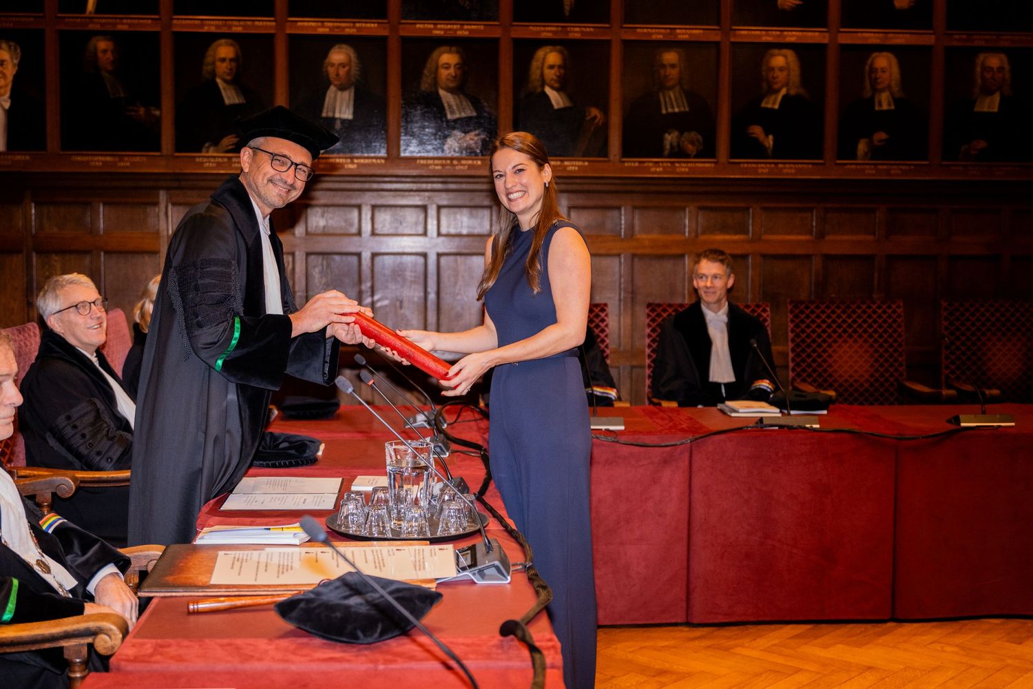 Graduate receiving diploma during formal ceremony in wood-paneled hall.