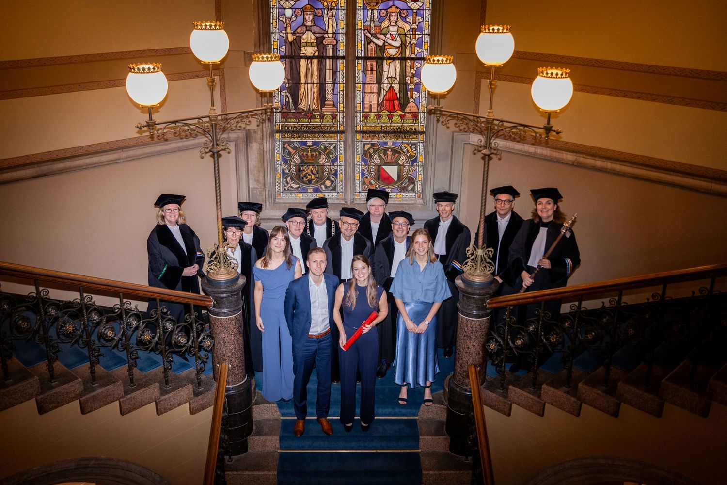Group photo of graduates and faculty on grand staircase with stained glass windows.