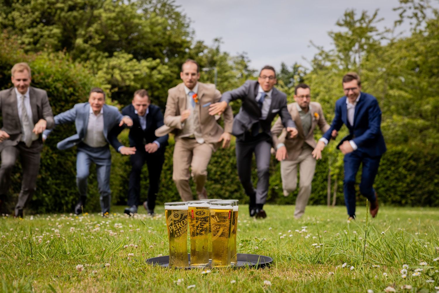 A group leaps joyfully towards three beer glasses on the grass during an outdoor celebration.