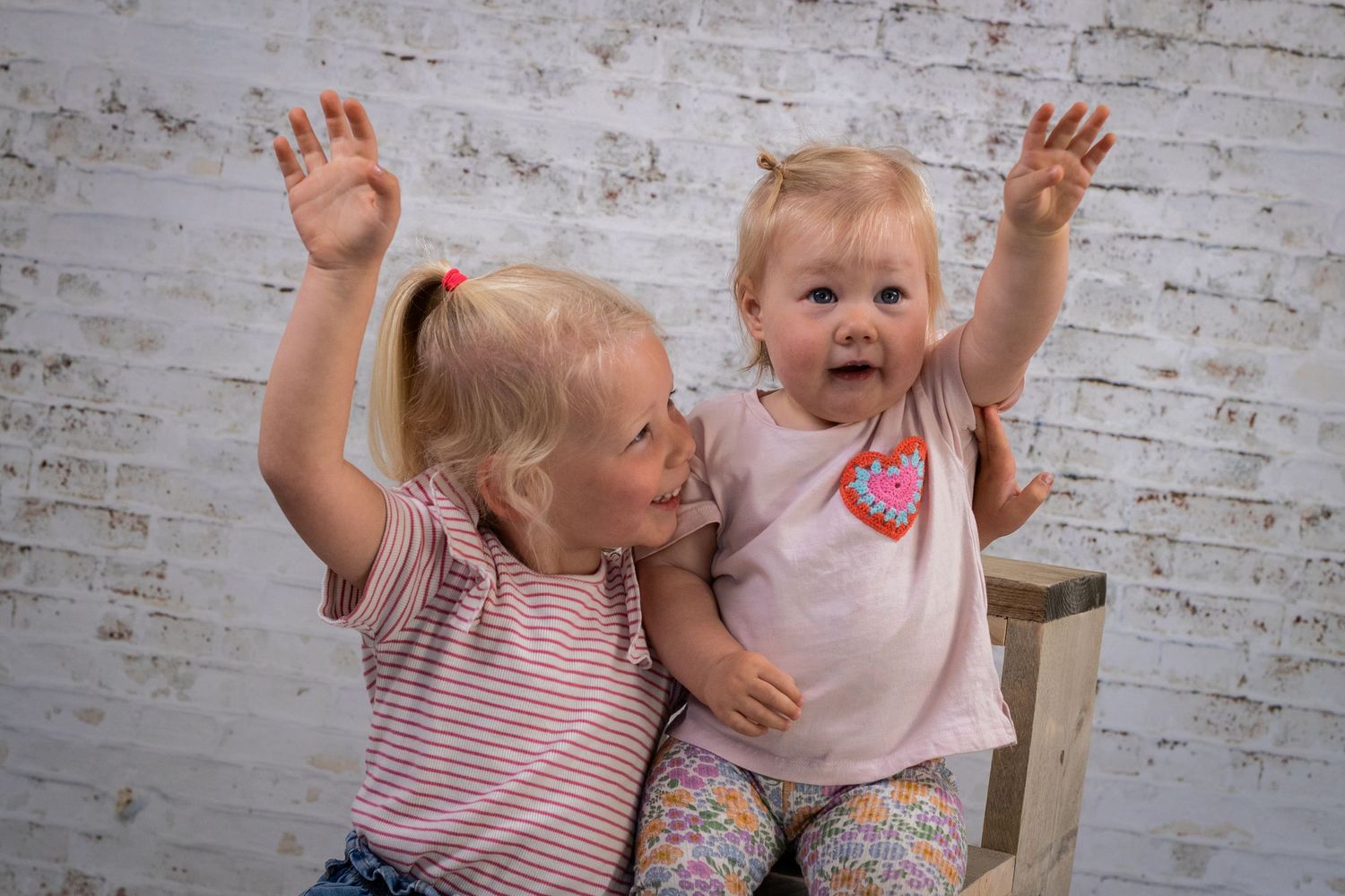 Young child wearing pink striped shirt playing and raising hands against white brick wall background.