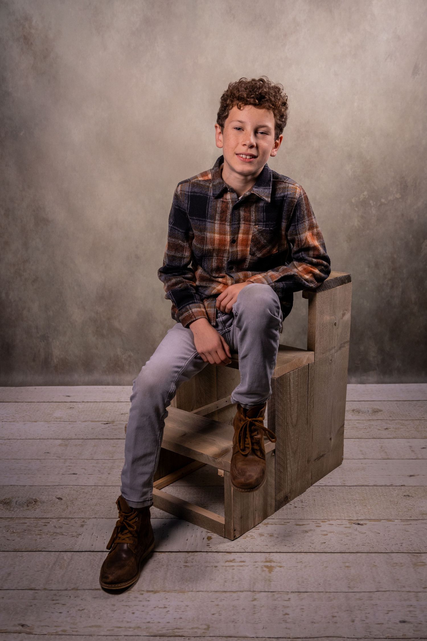 Young person in plaid shirt sits casually on wooden steps against gray backdrop for portrait session.