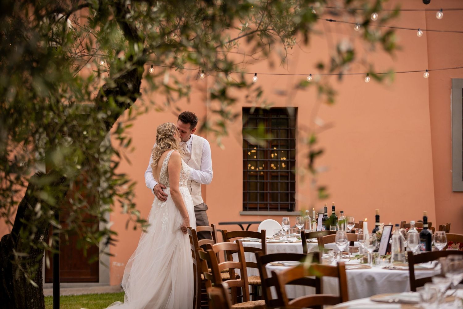 Elegant outdoor dining setup with white chairs at tables under string lights against a terracotta colored wall.