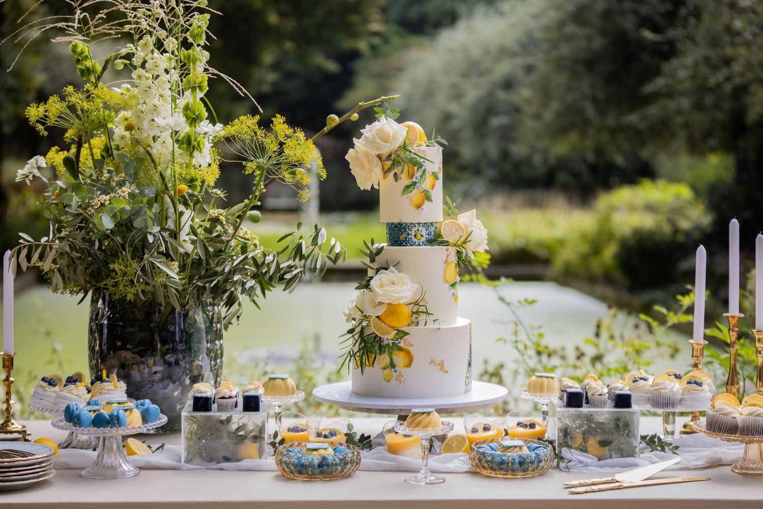Elegant outdoor wedding dessert table with tiered white cake, candles, and colorful pastries in a garden setting.
