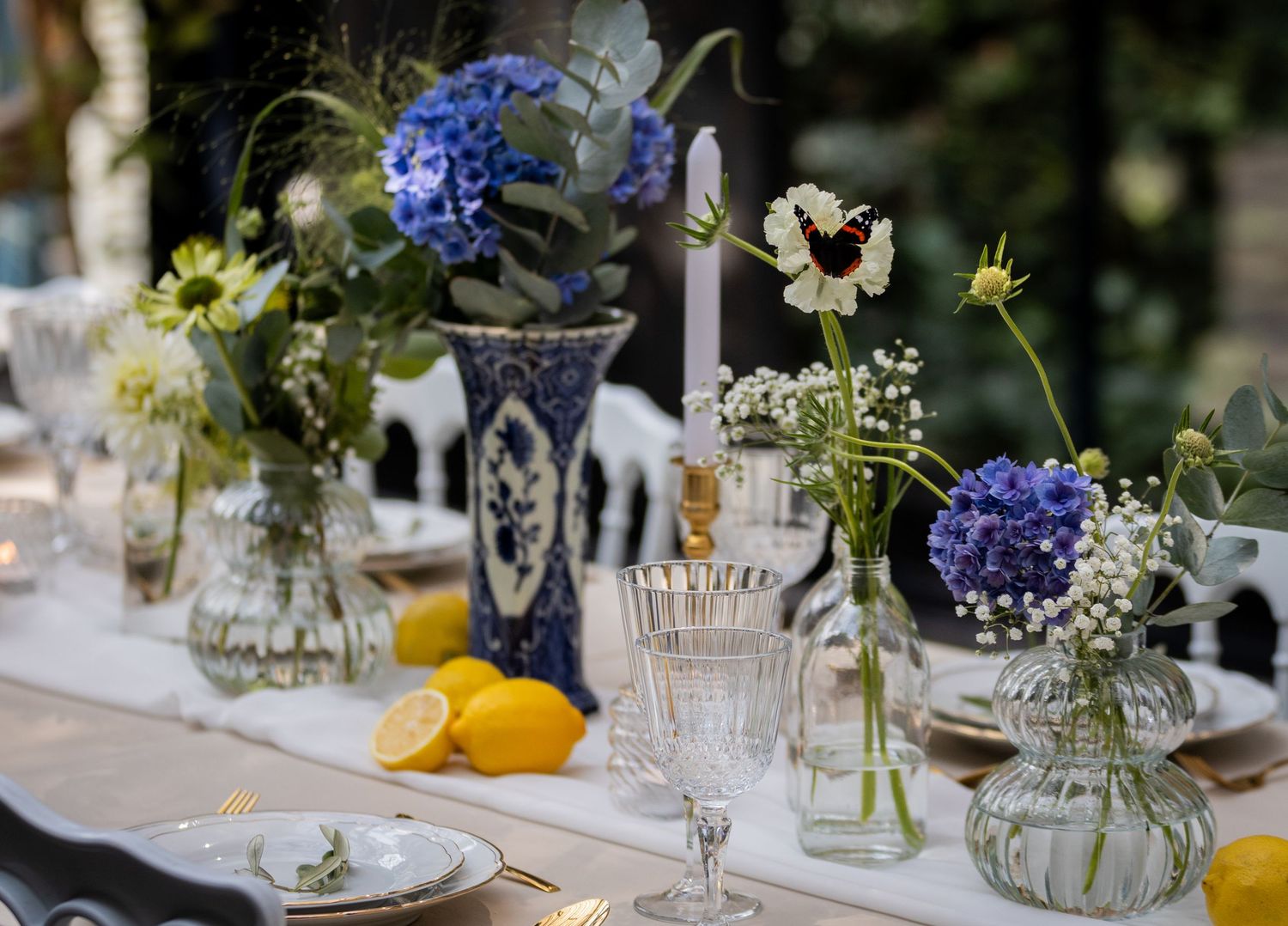 Table setting with glass vases of blue hydrangeas and white wildflowers alongside crystal glassware and fresh lemons.