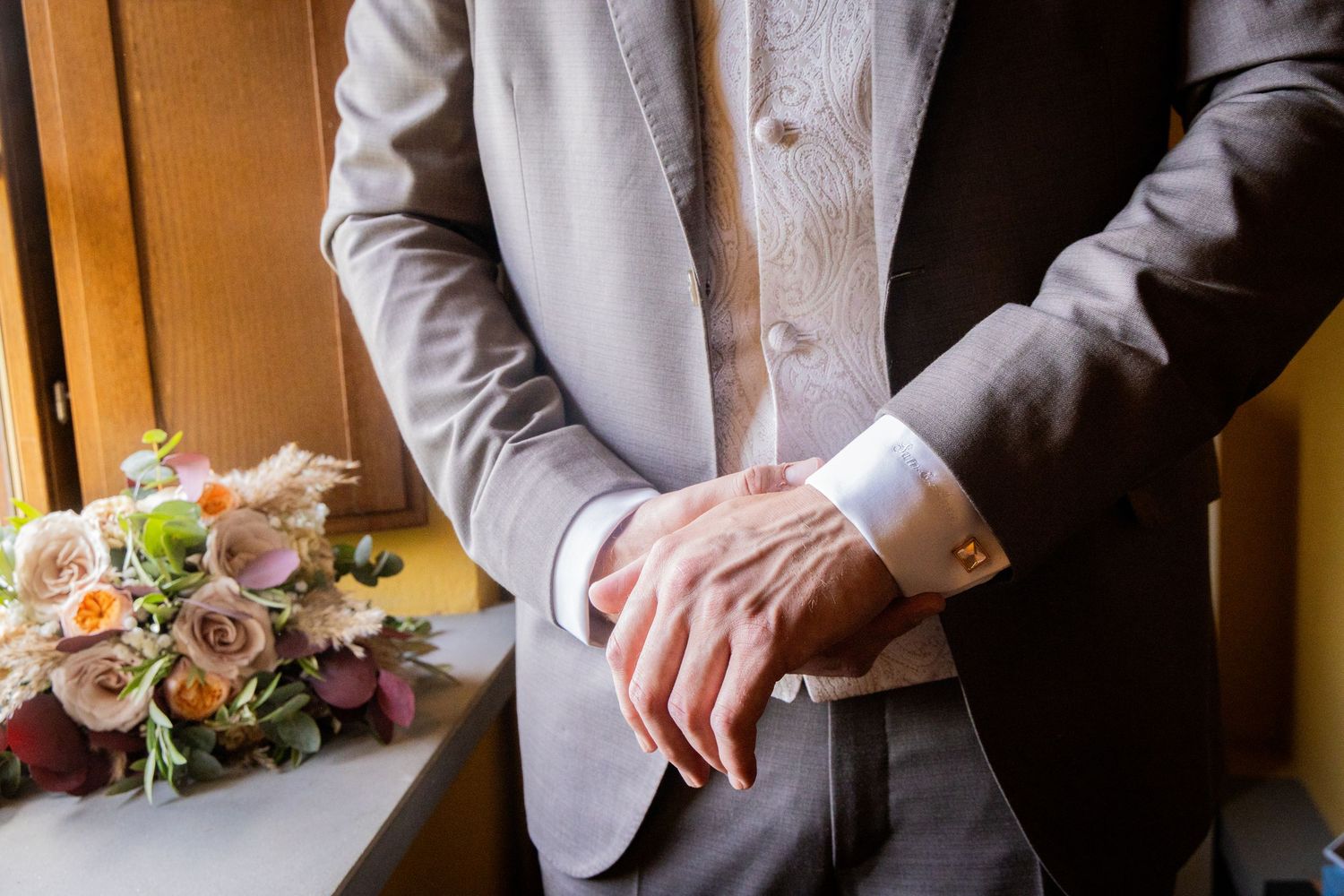 A groom in a gray suit jacket adjusts his cuff while a floral wedding bouquet rests nearby on a wooden surface.