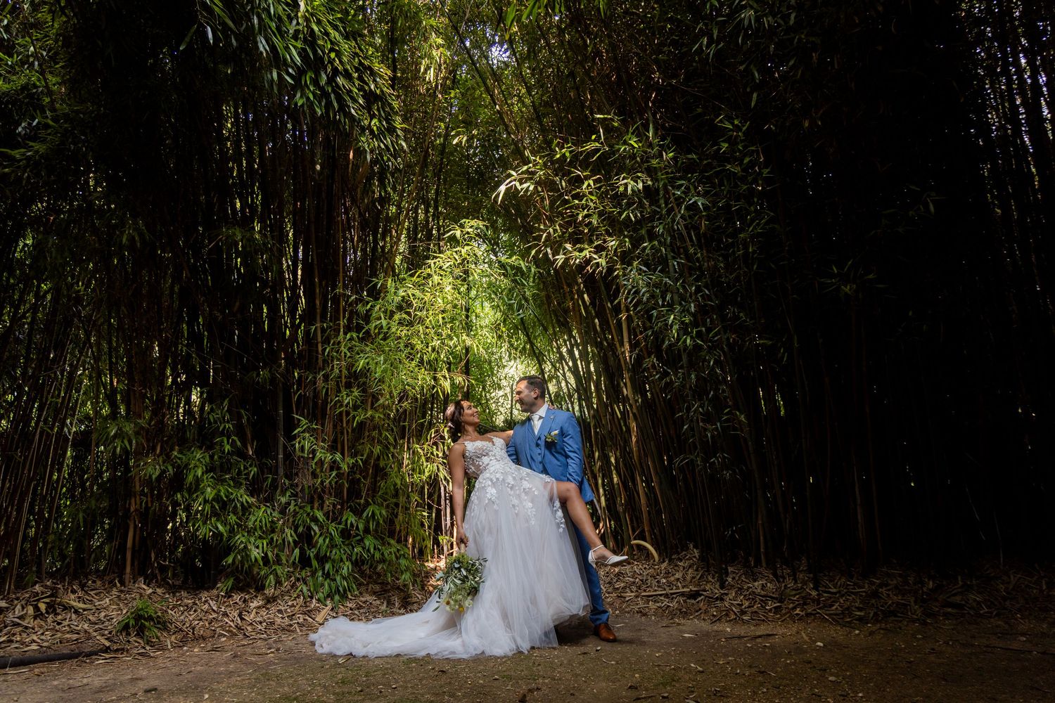 A couple poses romantically in a lush bamboo forest pathway while in formal wedding attire.