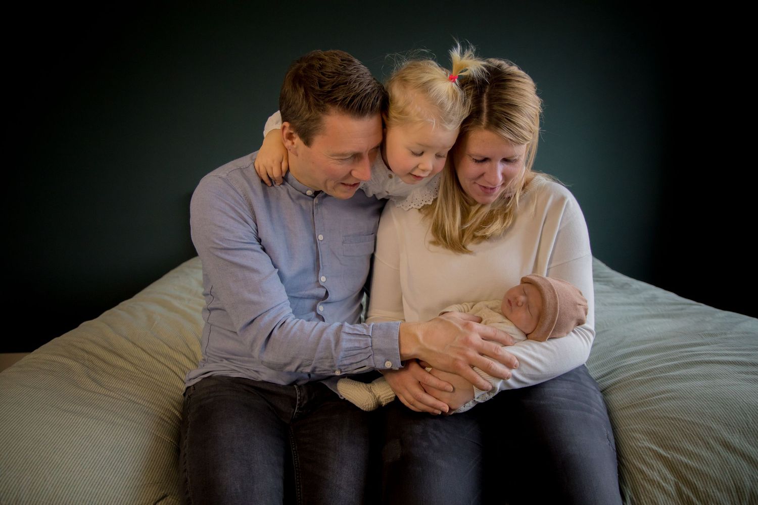 Family of three cuddling with newborn baby on bed against dark backdrop.