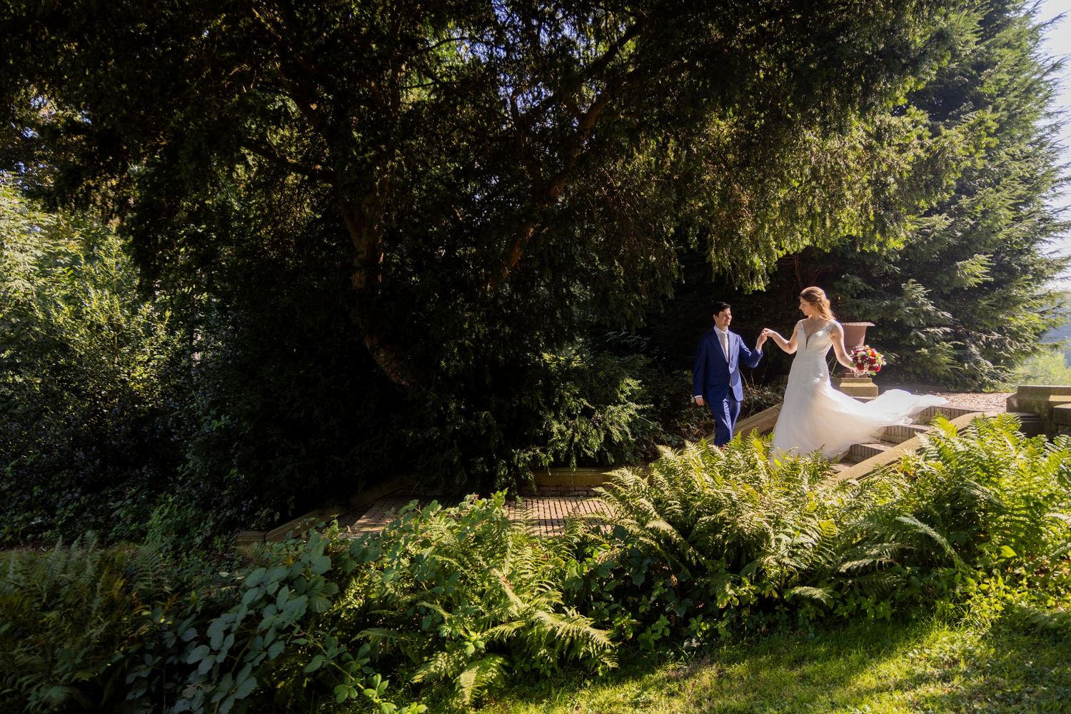 Wedding couple walks along garden path surrounded by lush greenery.