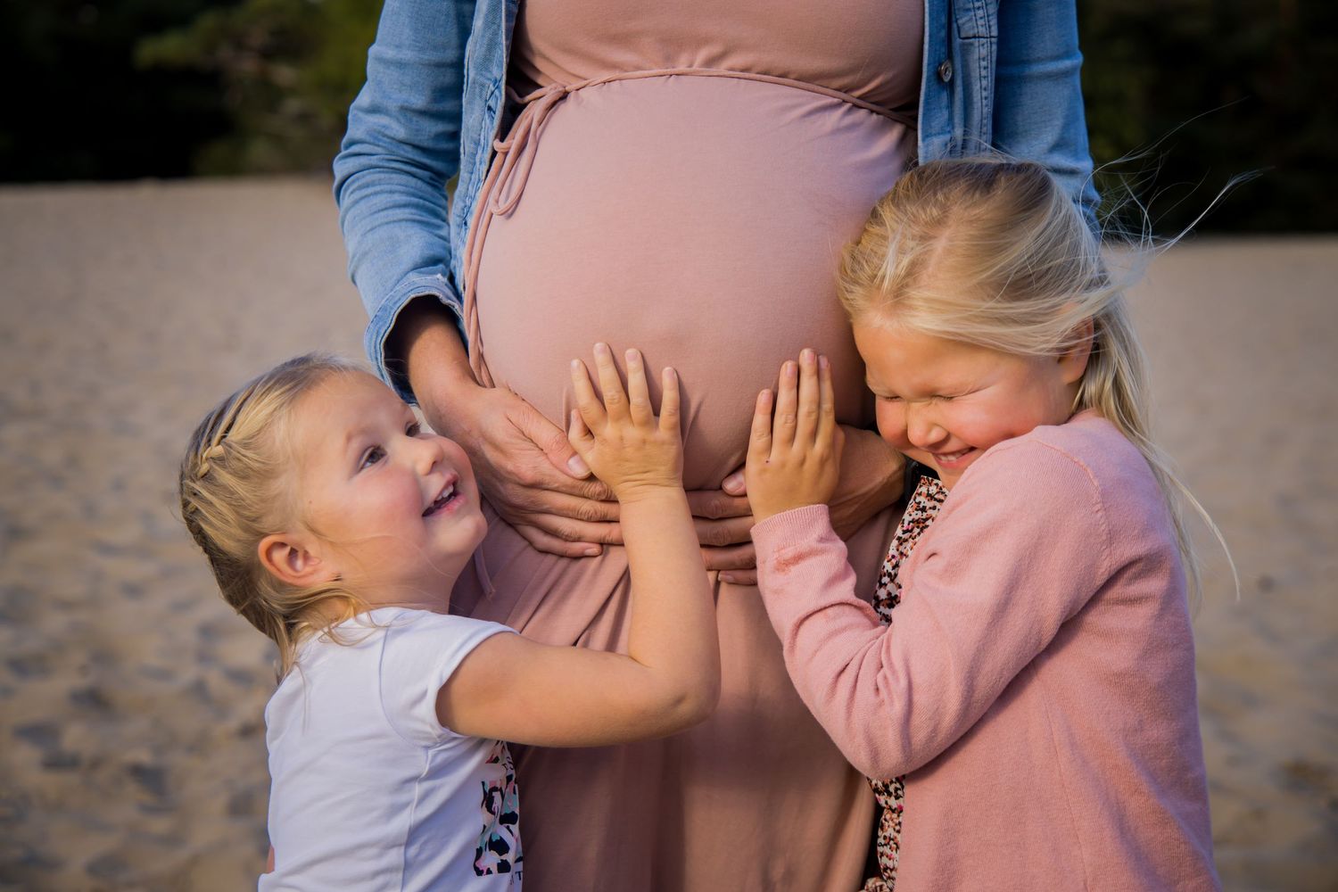 Two small children touch an expectant mother's belly on a beach at sunset.