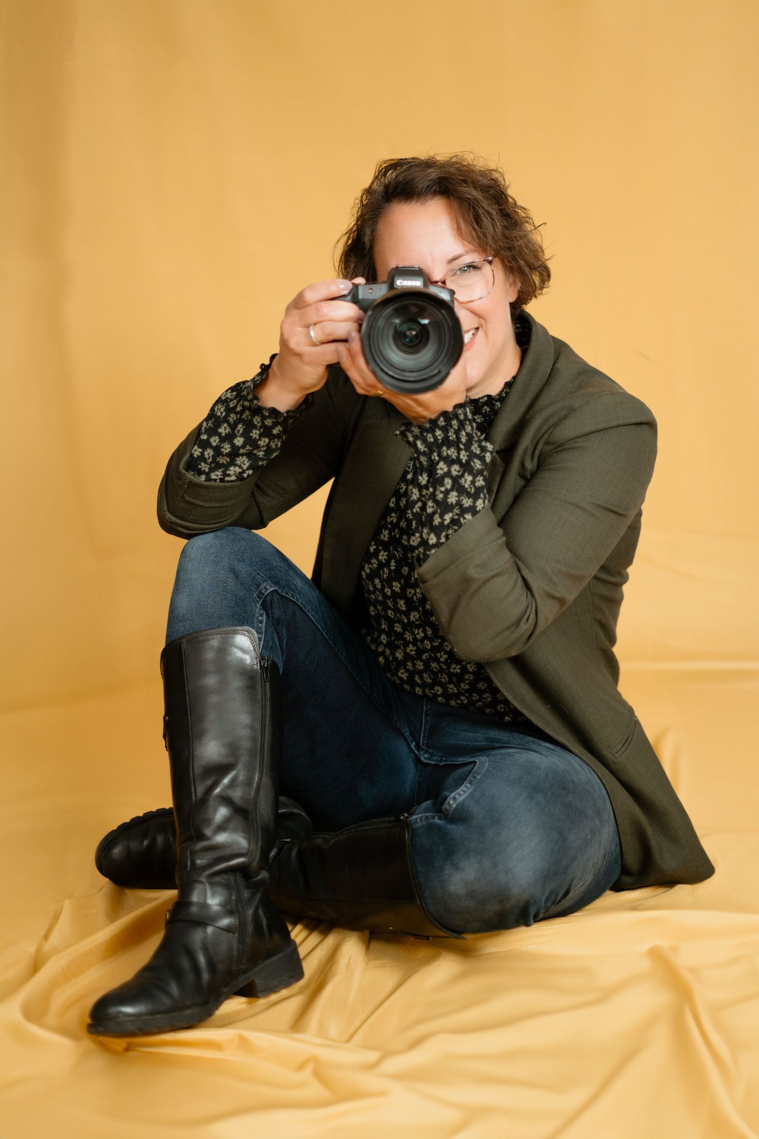 Camera photographer sitting on yellow backdrop wearing green jacket, patterned shirt and knee-high boots.