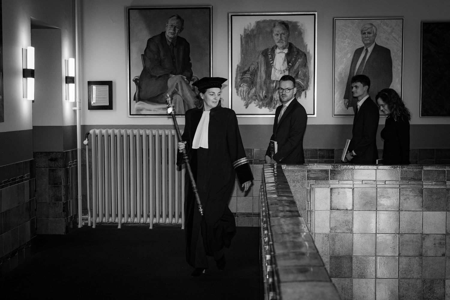 Black and white image of a formal gathering beneath vintage portraits in an institutional hallway.