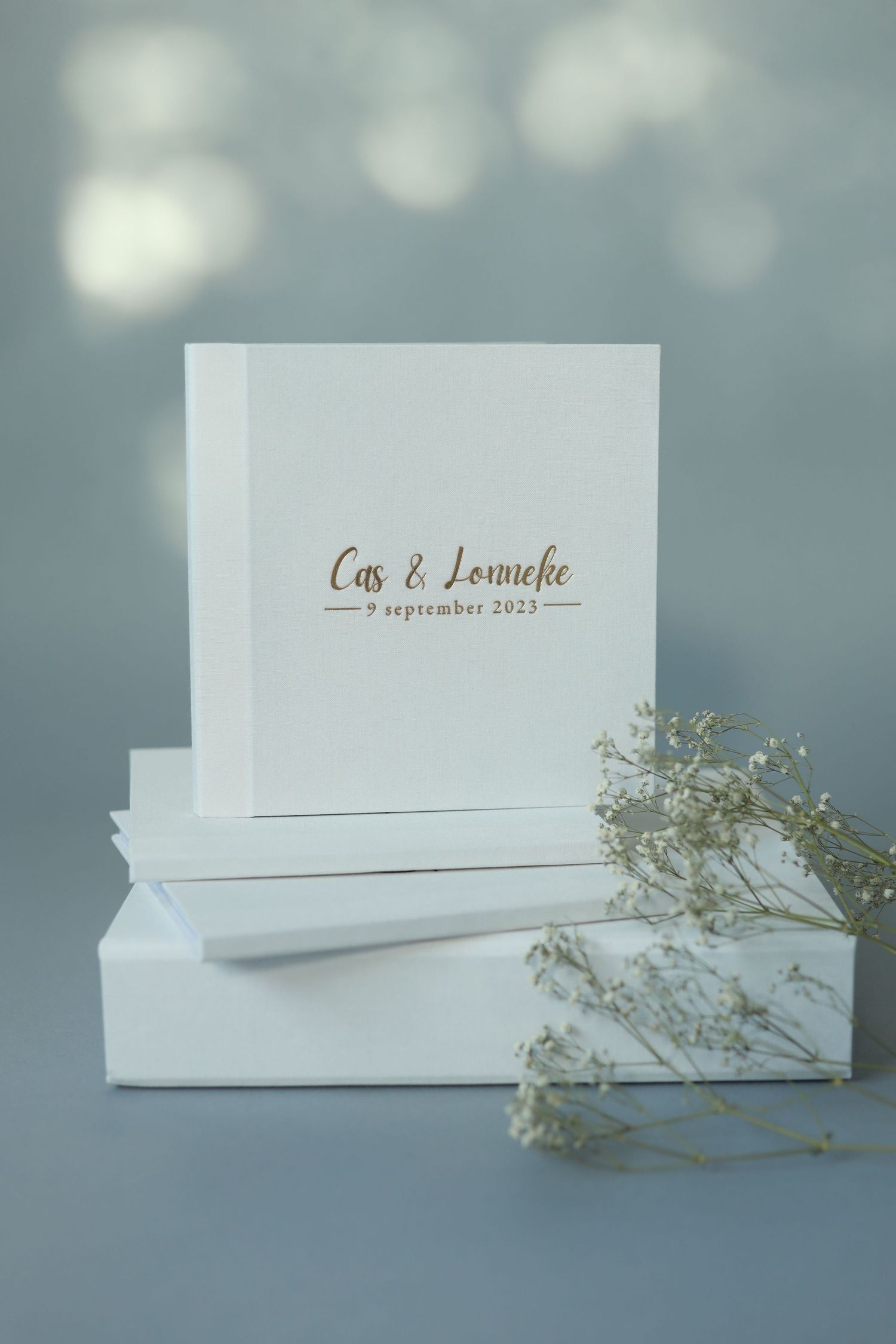 Elegant white wedding album with gold lettering and baby's breath floral decoration against a soft gray background.