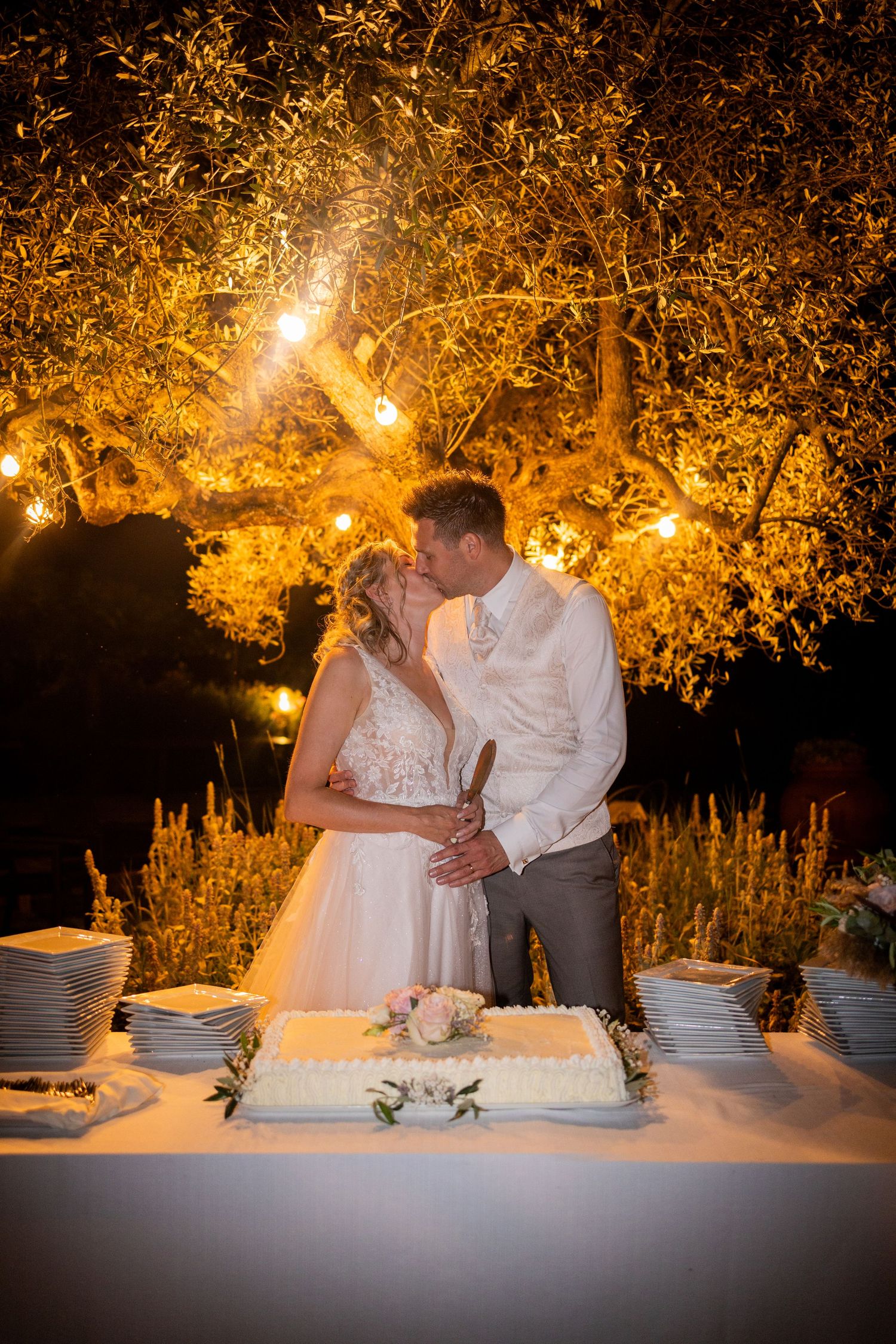 A romantic outdoor evening wedding reception with a couple standing by their wedding cake under illuminated trees.