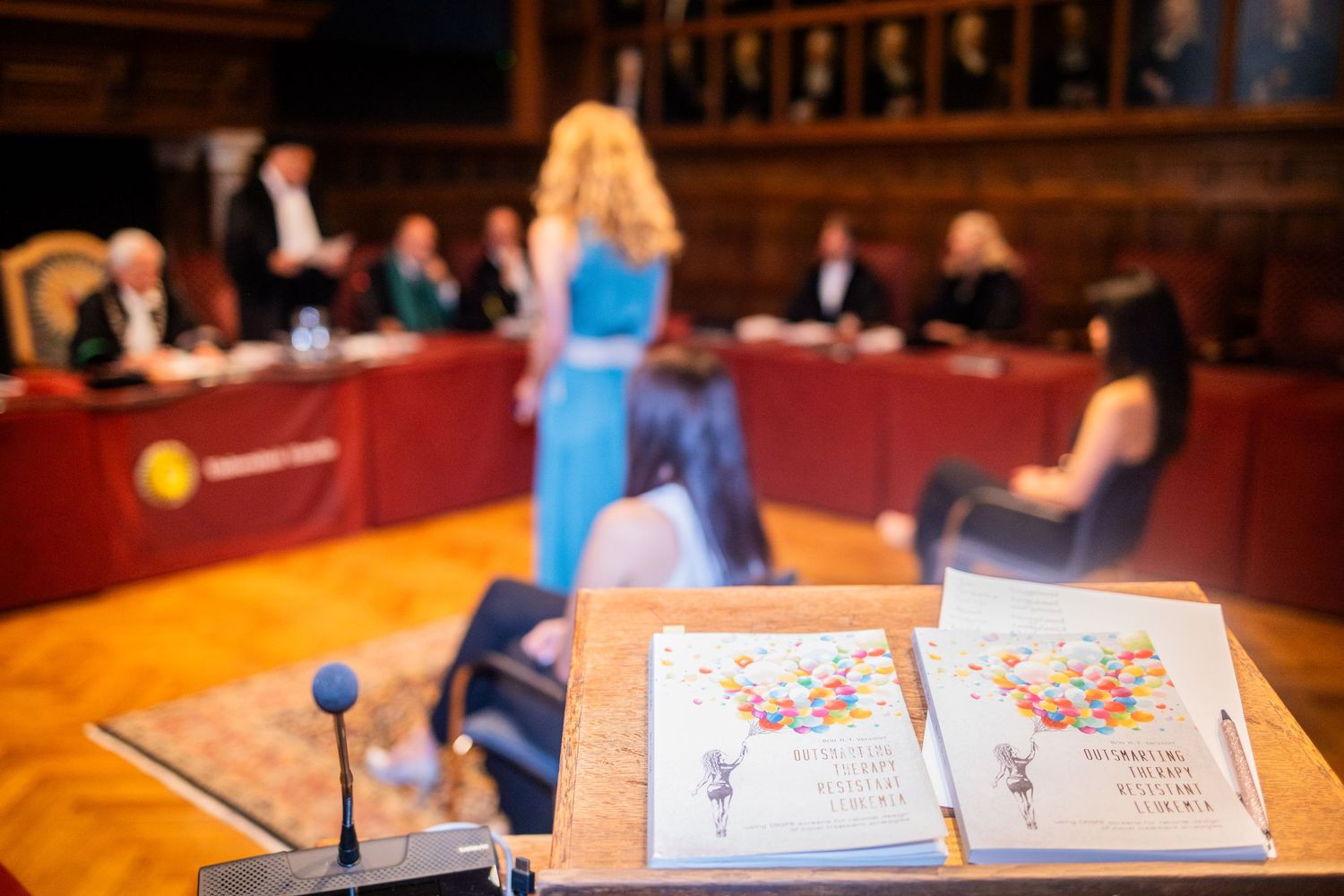 Academic ceremony books displayed on table with audience in background.
