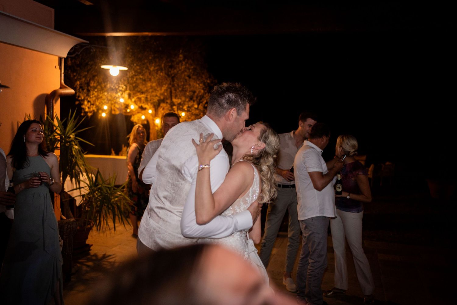 A couple shares a romantic dance moment outdoors at an evening reception with warm lighting and other dancers in background.