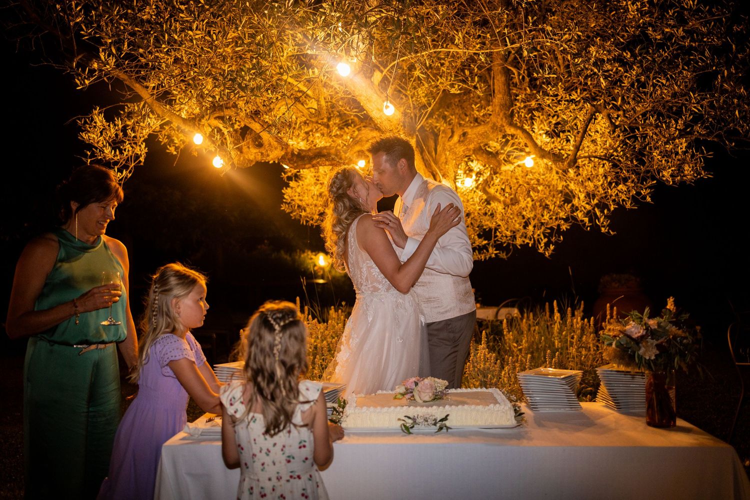 Newlywed couple cutting their wedding cake at night under illuminated trees with young flower girls watching.