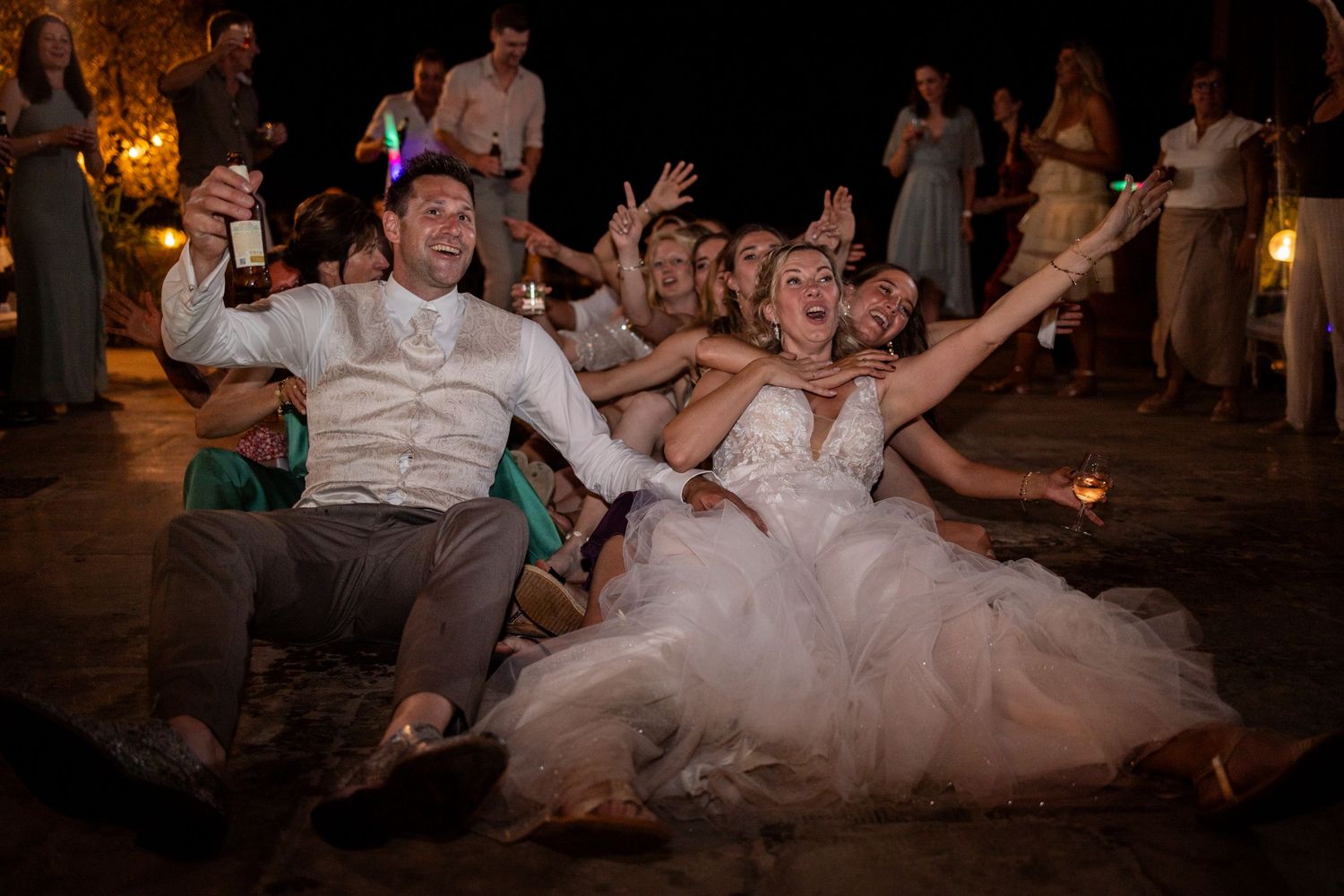 Joyful newlyweds and friends dancing on the ground at a nighttime wedding reception with ambient lighting.