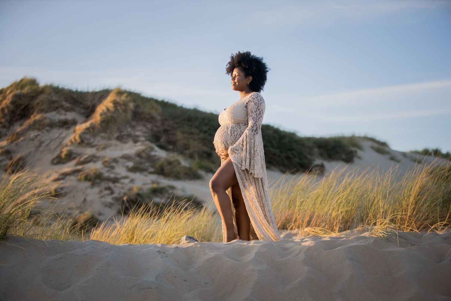 Ethereal figure in white dress stands among beach dunes and sea grass at golden hour.