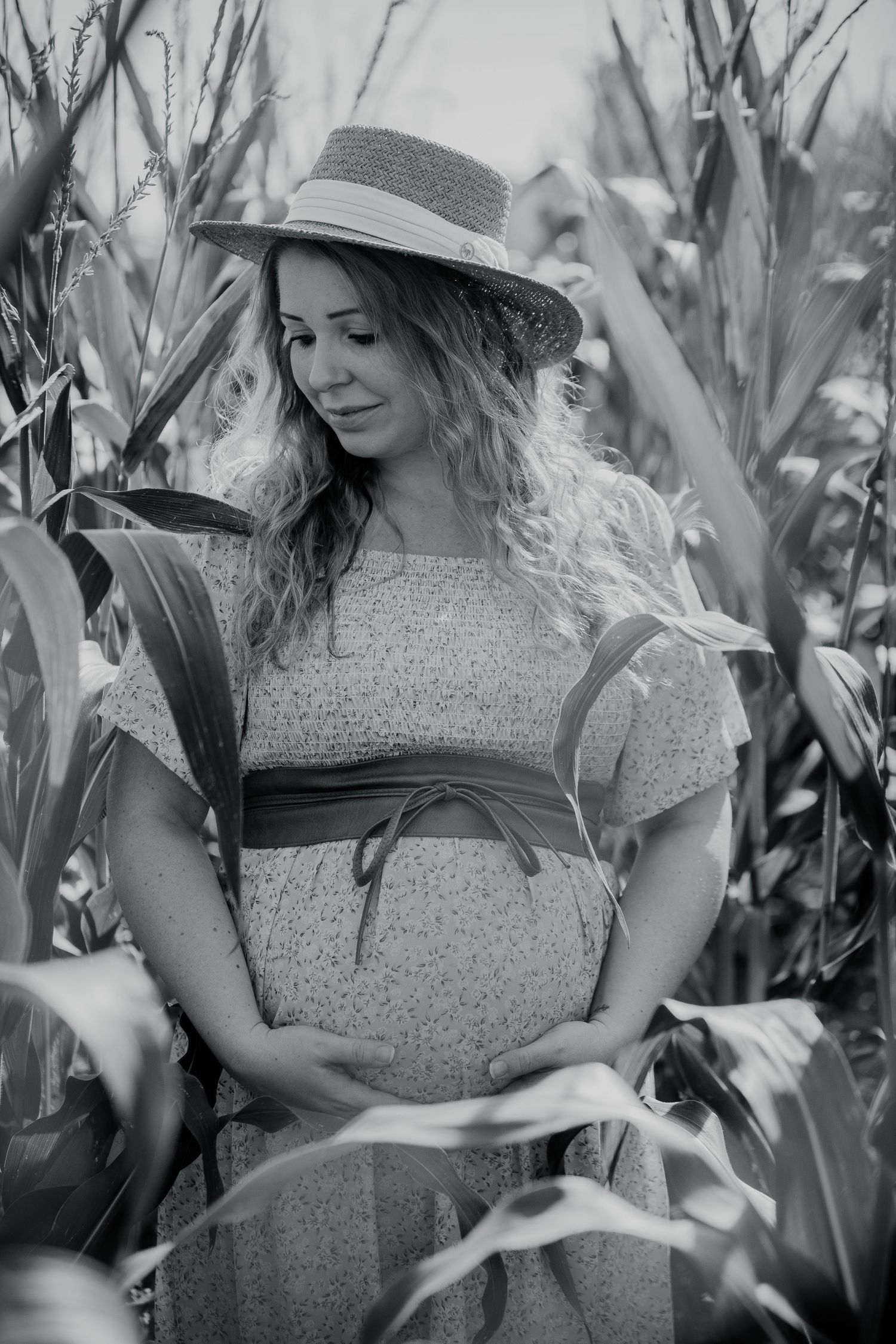 Woman in straw hat posing thoughtfully among tall corn stalks in black and white.