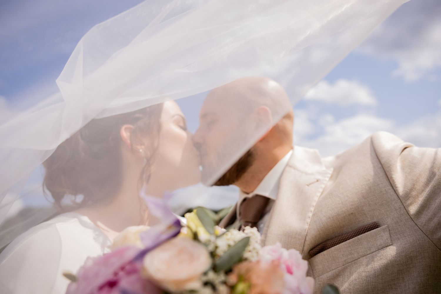 Close-up of romantic moment under flowing white veil with pink flowers in foreground.