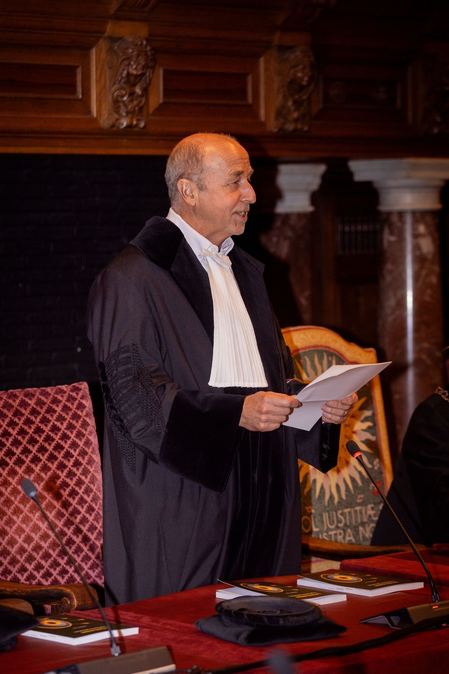A distinguished speaker in academic robes addresses an audience from behind a podium.
