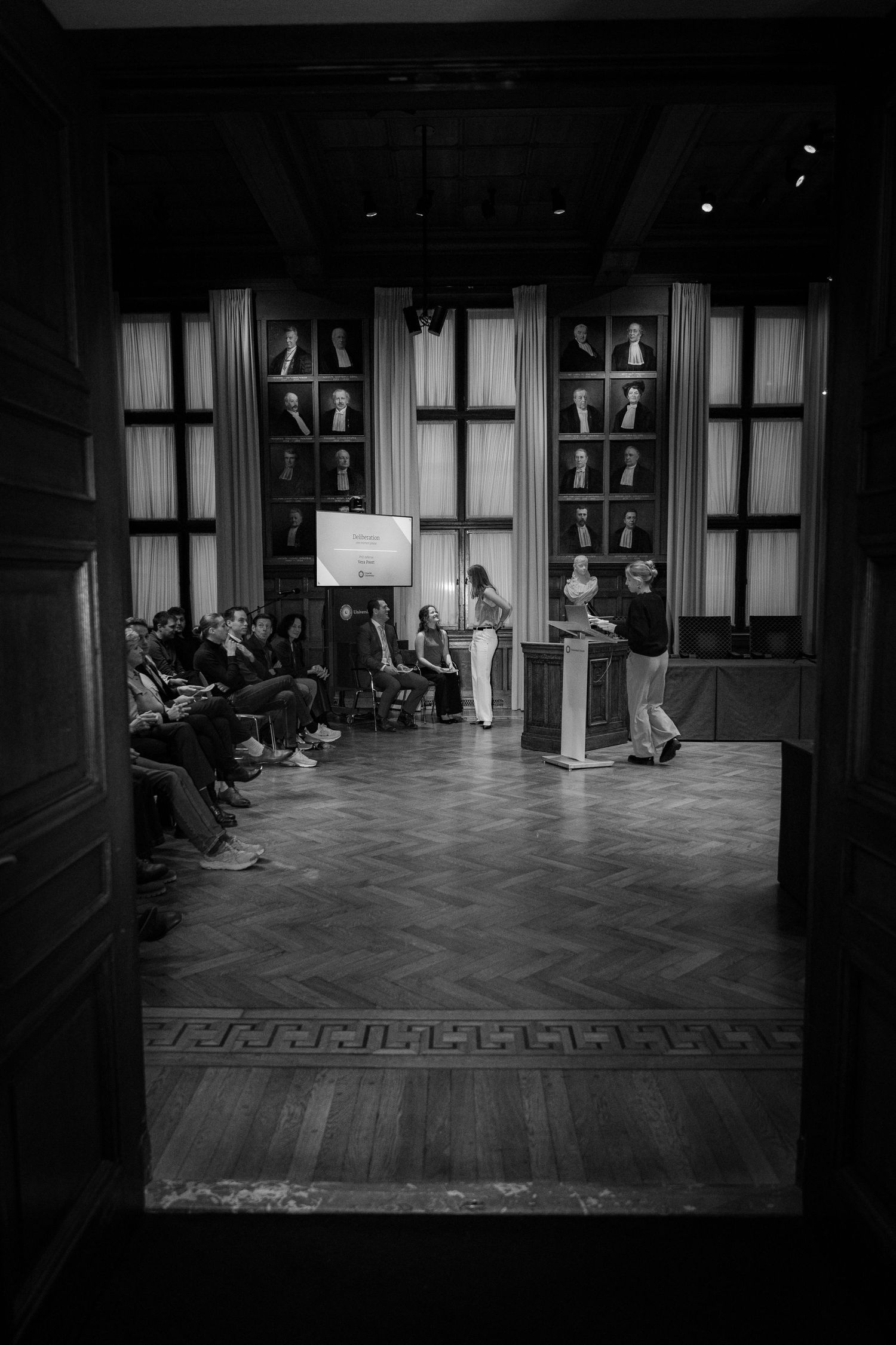 Black and white photograph of a formal academic presentation in an elegant room with ornate windows and wooden flooring.