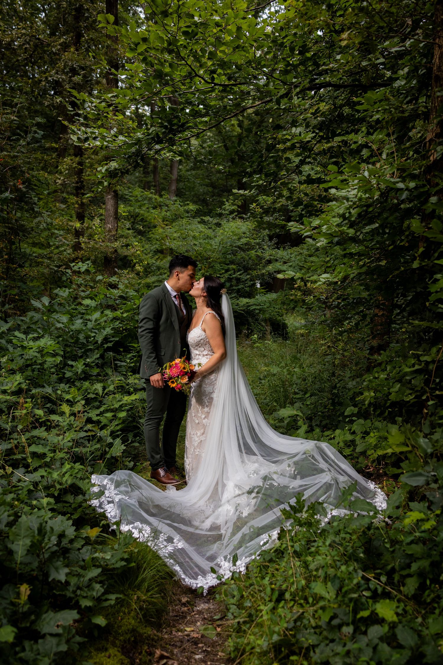 Wedding couple poses in lush forest setting with dramatic flowing veil and autumn bouquet.
