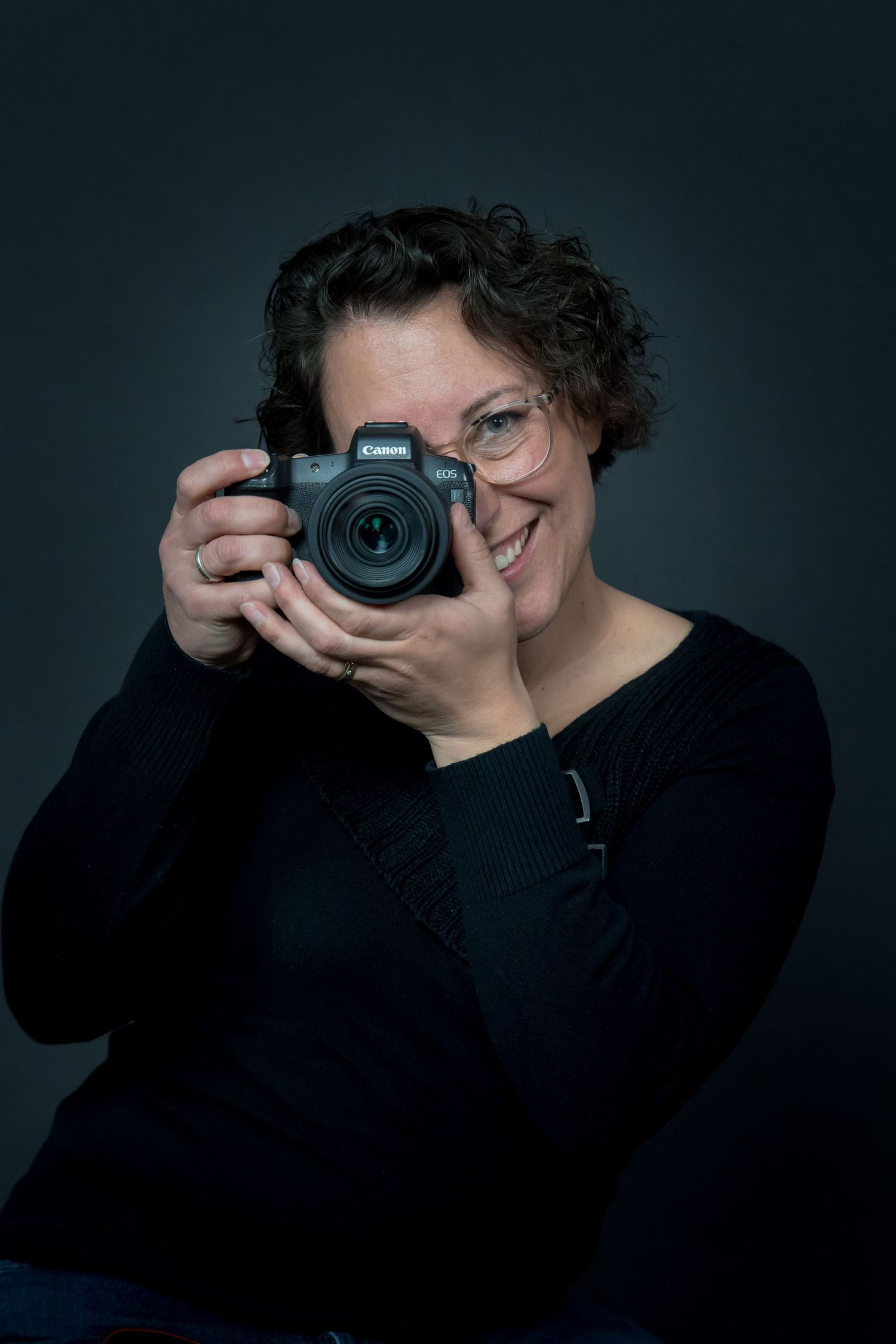 A photographer smiles while holding a professional DSLR camera against a dark background.