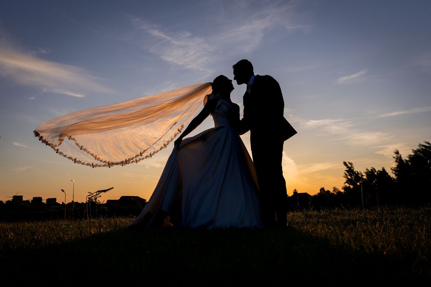 Silhouettes of newlyweds pose against a dramatic sunset sky with flowing veil.