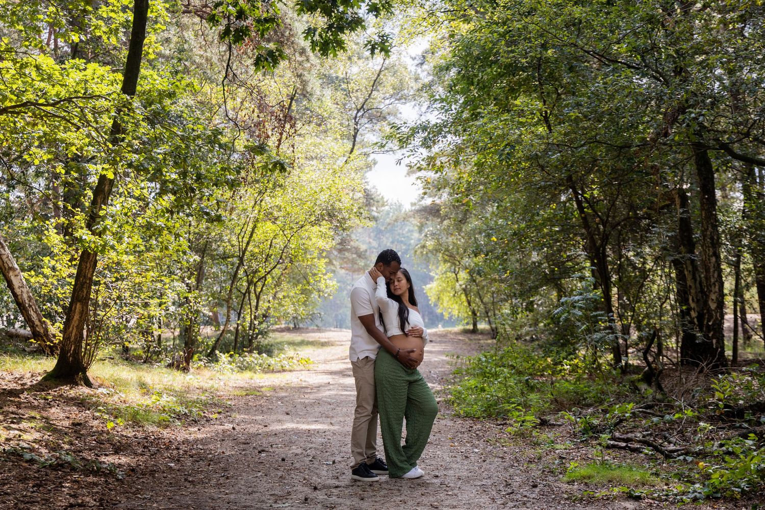 Couple embracing on a sunlit forest path surrounded by lush green trees.