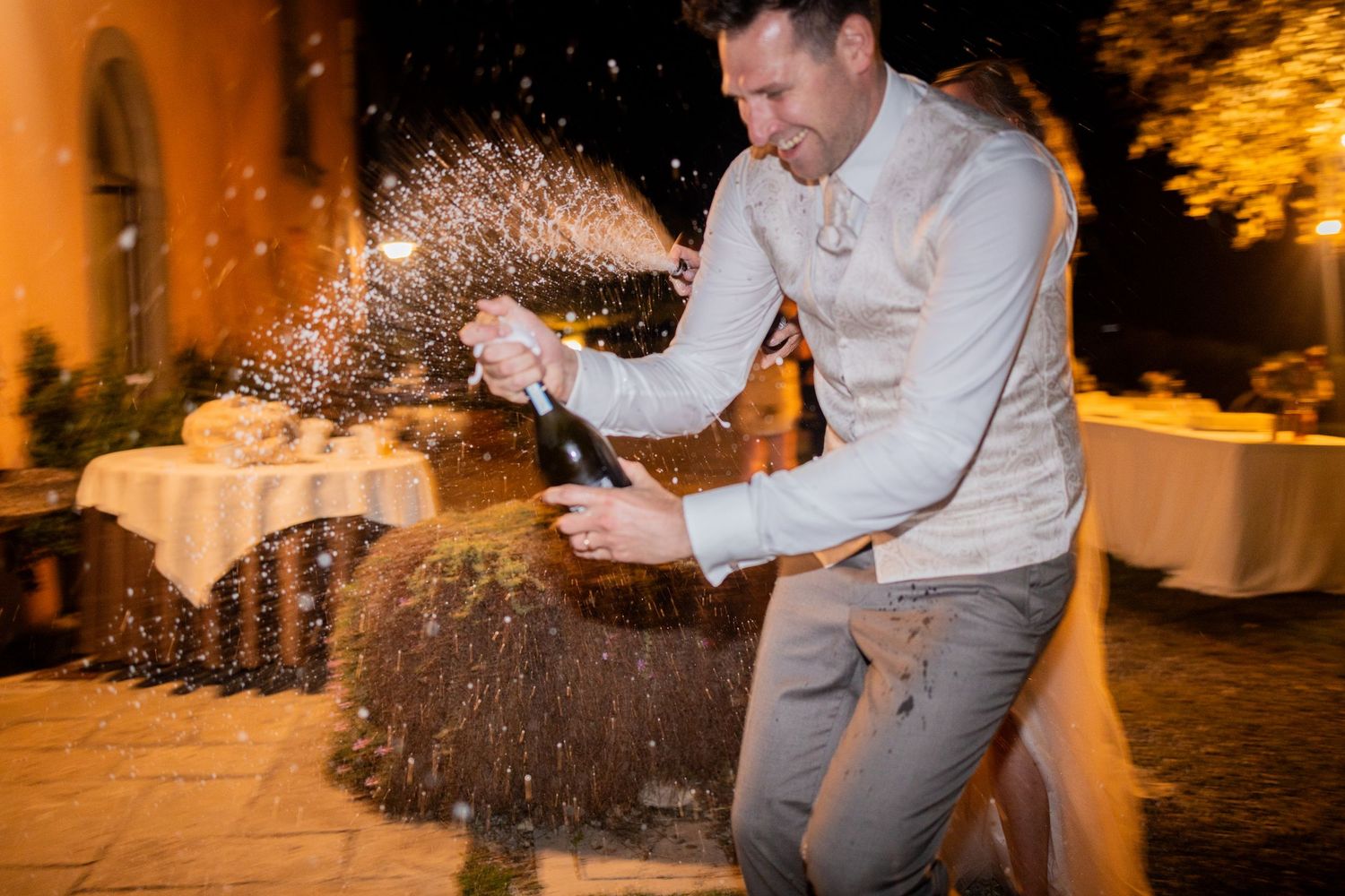 Person in a gray suit and white shirt sprays champagne outdoors at a nighttime celebration event, creating a dramatic splash.