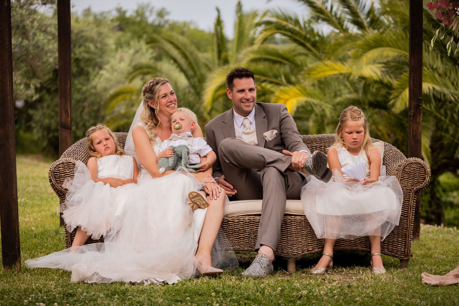 Wedding party relaxes on an elegant brown couch outdoors surrounded by lush palm trees and greenery.