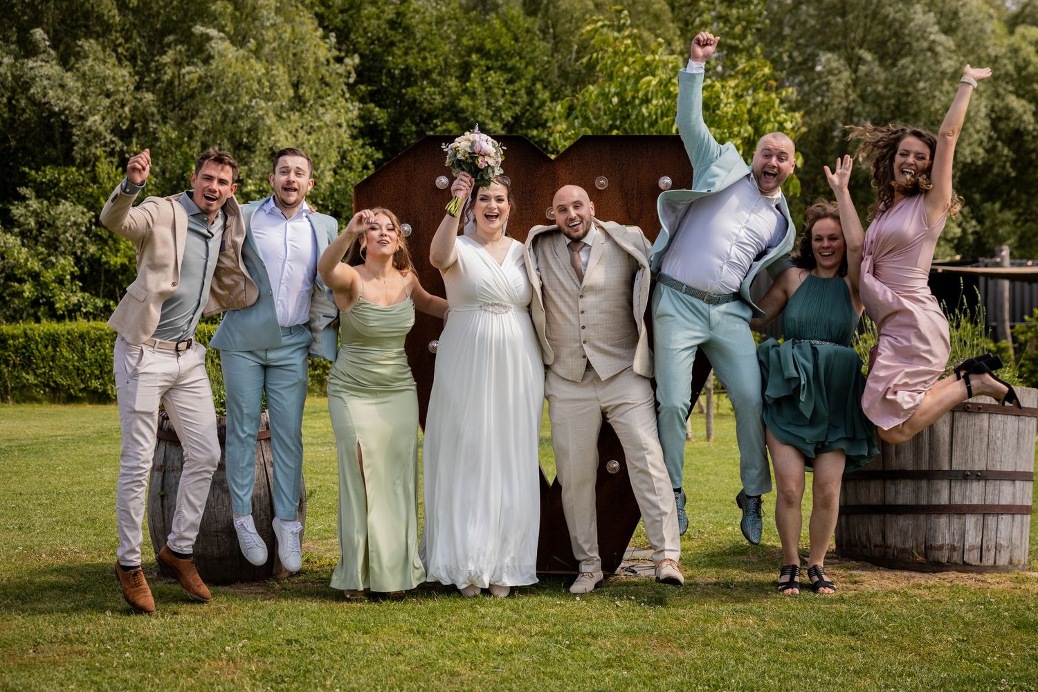 Wedding party jumps joyfully for a celebratory group photo in a sunny outdoor garden setting.