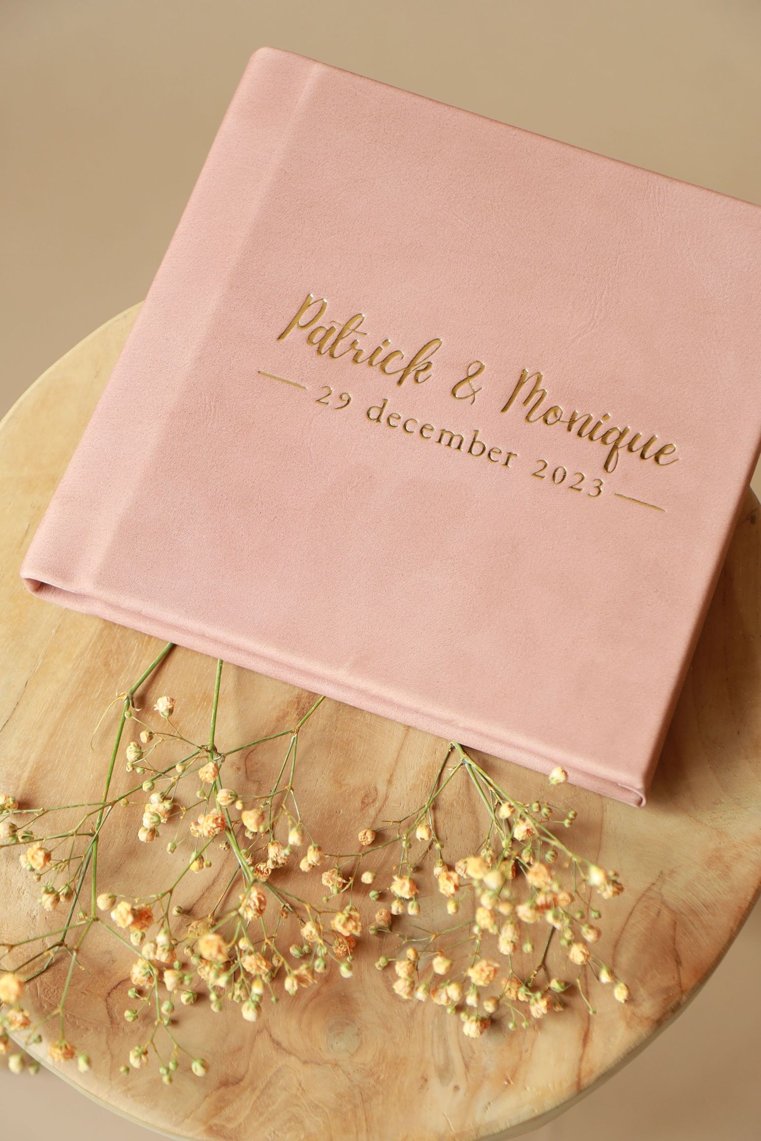 Pink wedding album with 'Patrick & Monique' and date in gold text, displayed with delicate baby's breath flowers on wooden surface.