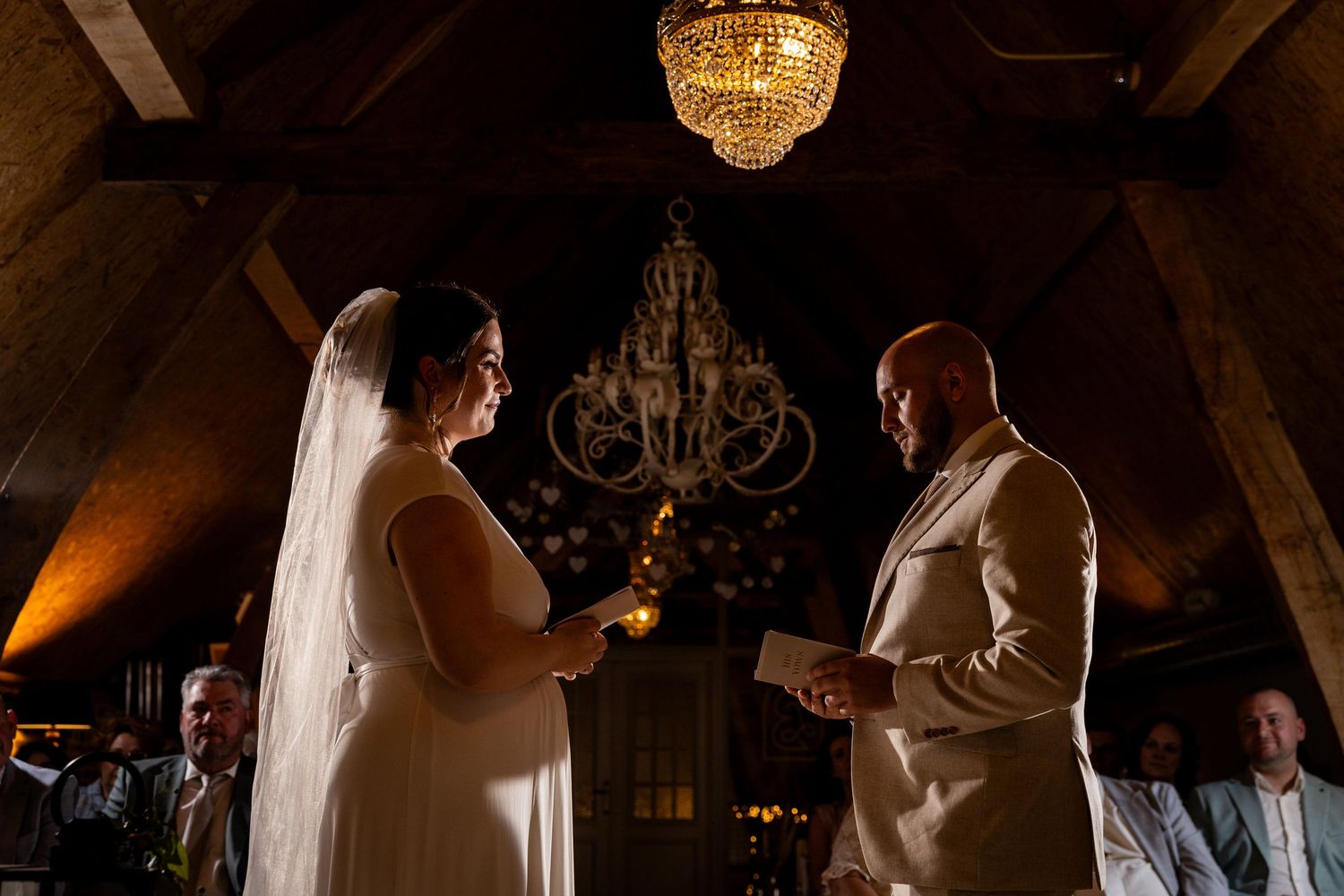 Wedding ceremony in rustic wooden venue with crystal chandelier and dramatic lighting.