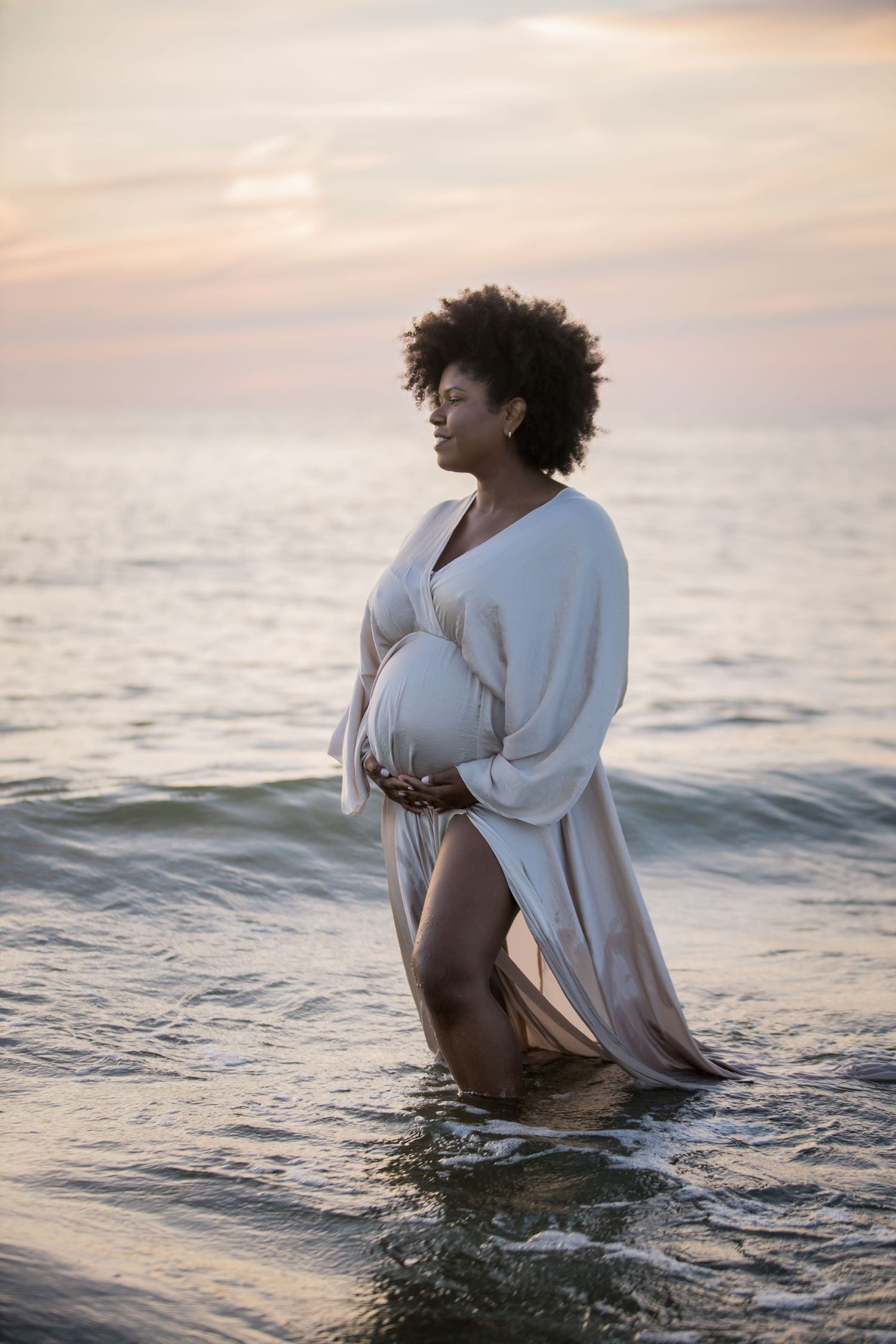 Expecting mother in white dress stands in ocean waves during sunset.