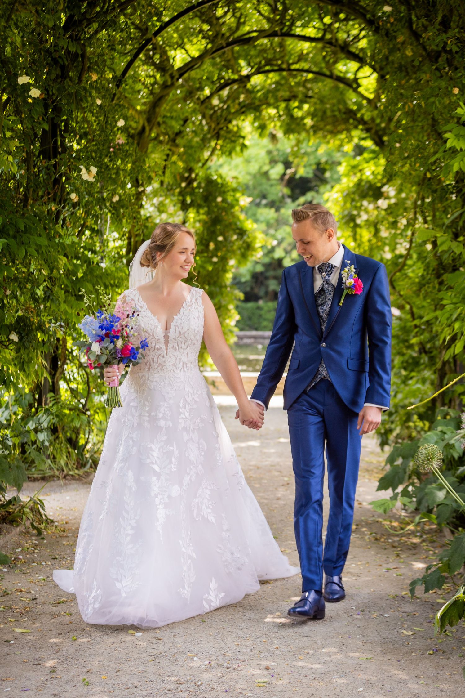 A couple in wedding attire walk hand-in-hand through a lush garden archway on their special day.