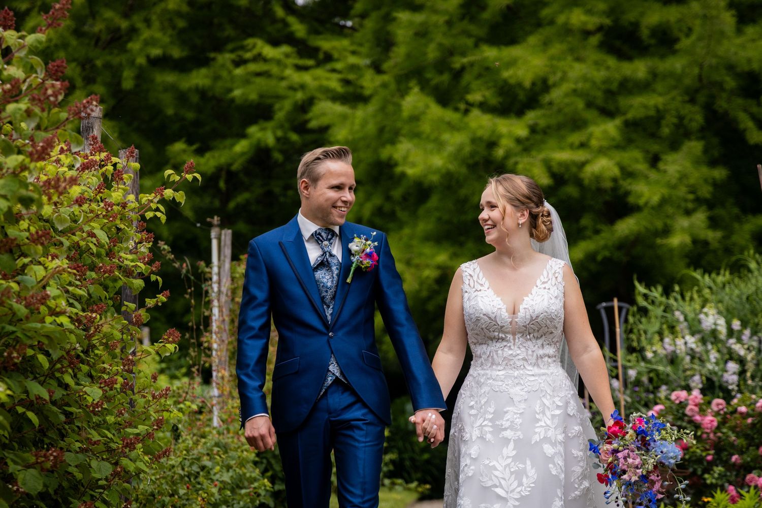 Newlyweds in blue suit and white dress walking hand in hand through a lush garden setting with purple flowers.