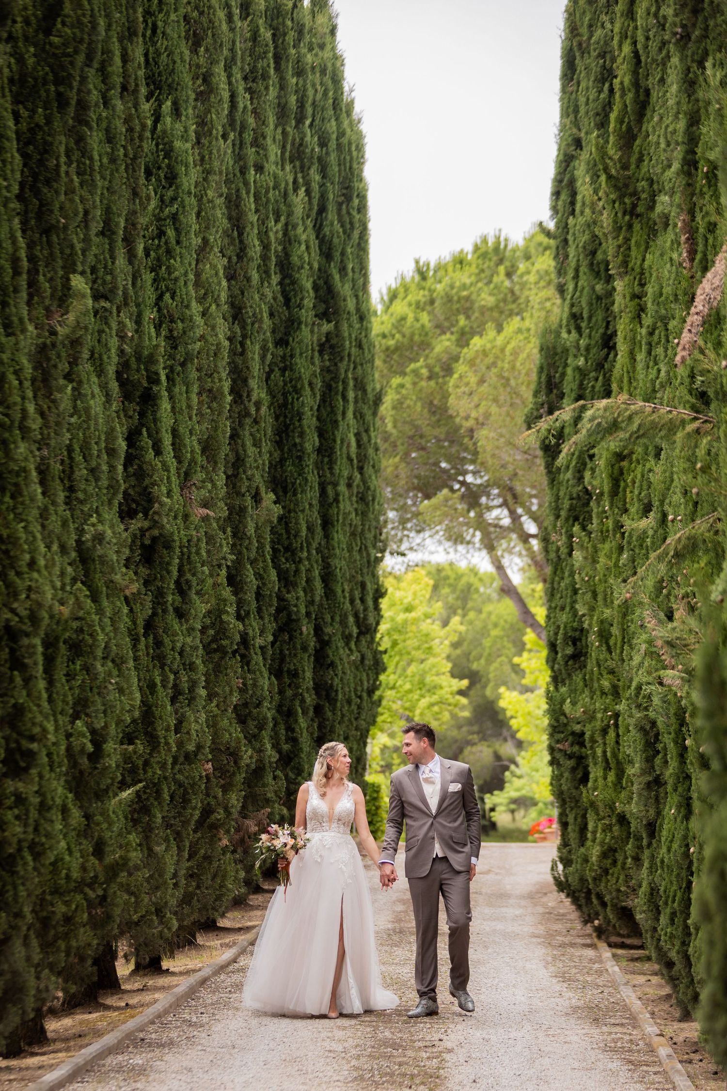 A couple walks down a garden path lined with tall cypress trees in a romantic Mediterranean setting.