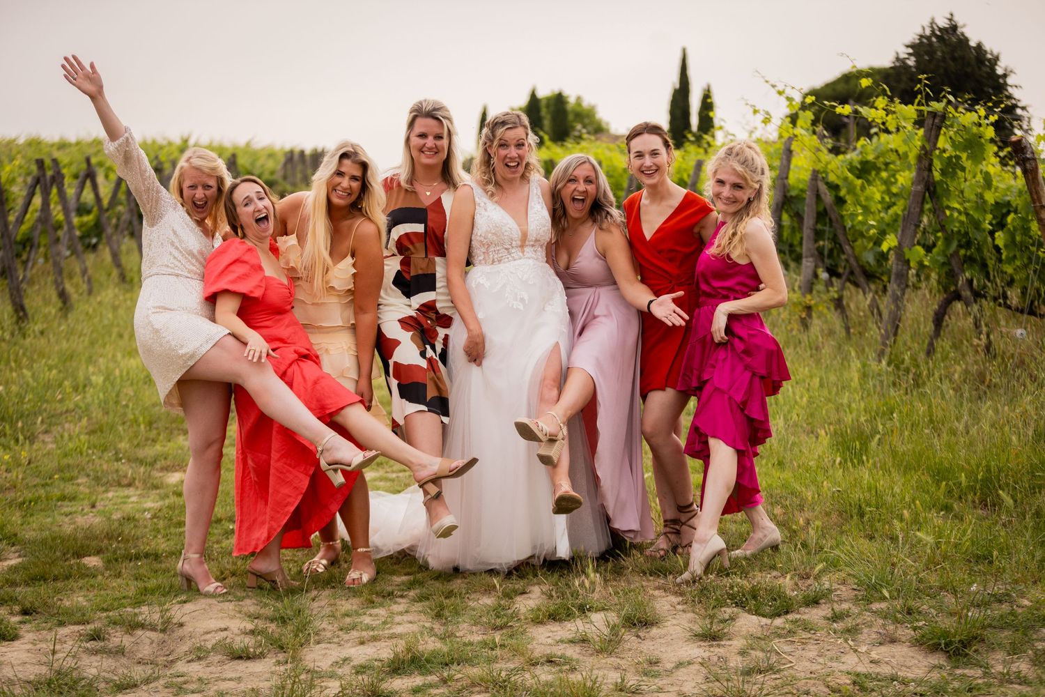 A celebratory group in colorful dresses poses joyfully together in a vineyard setting at sunset.