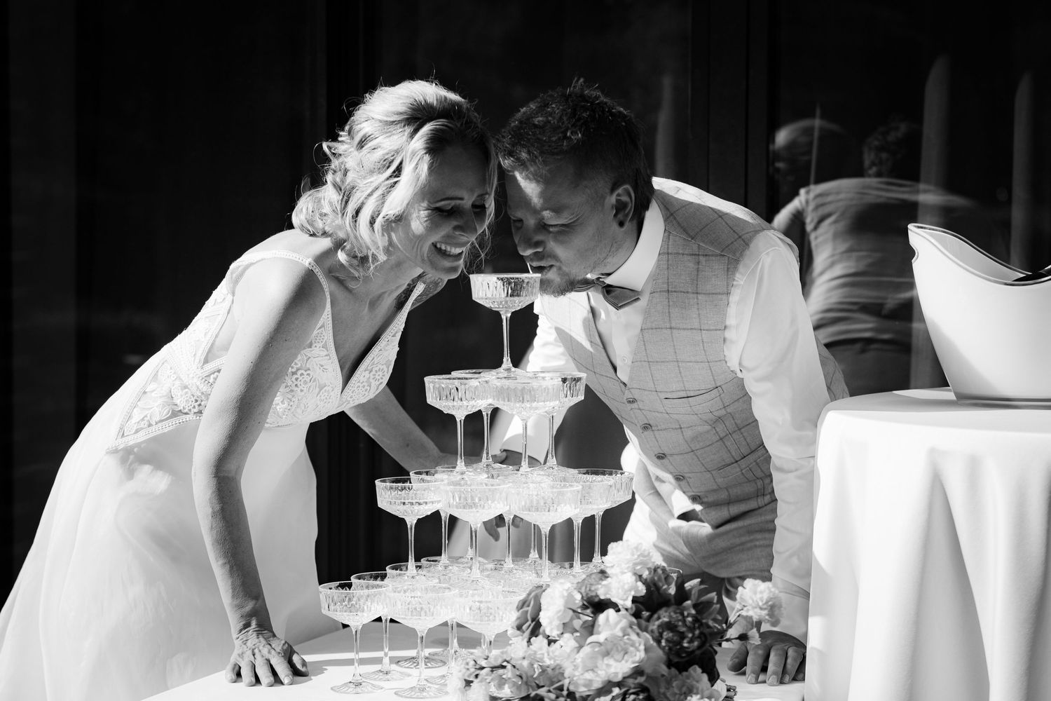 Black and white photo of newlyweds leaning over a champagne tower at their wedding reception.