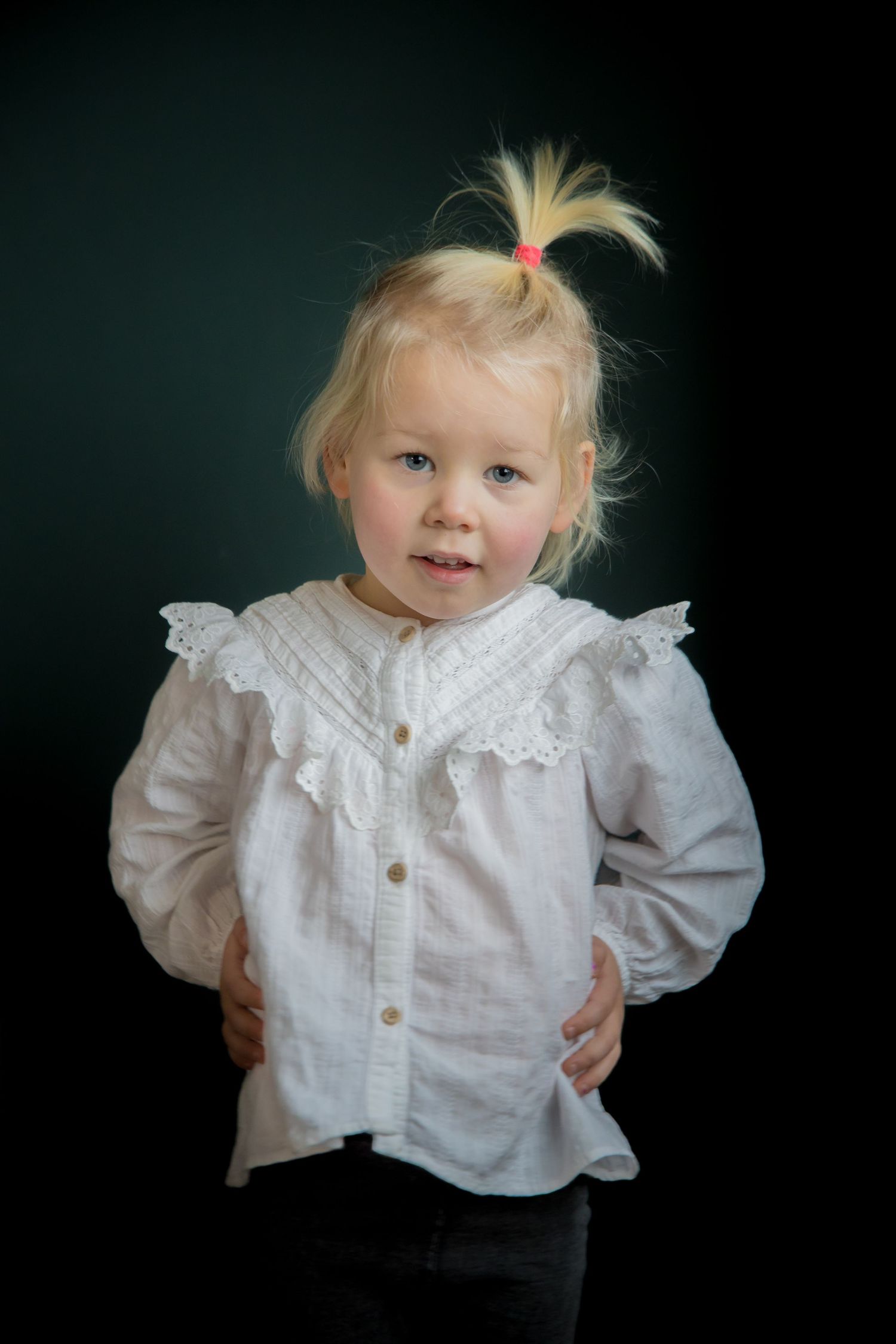 Young child in white ruffled shirt with blonde hair ponytail against dark background.