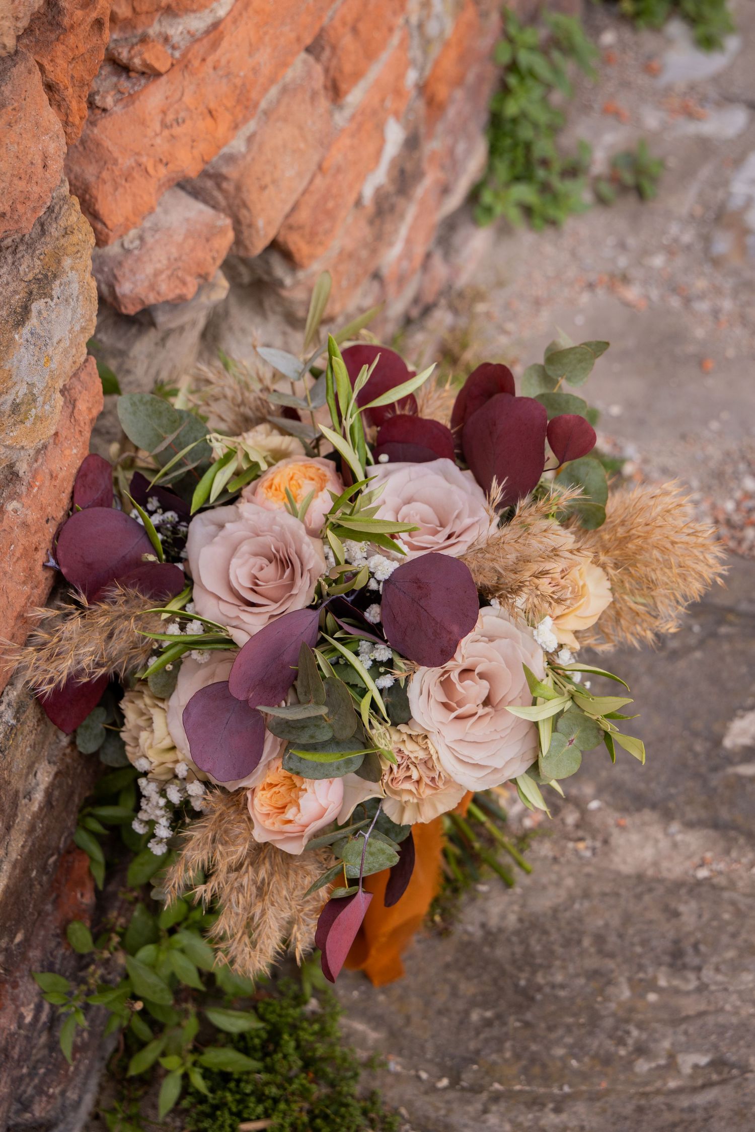 Elegant wedding bouquet with blush roses, burgundy eucalyptus, and dried pampas grass against rustic brick wall.