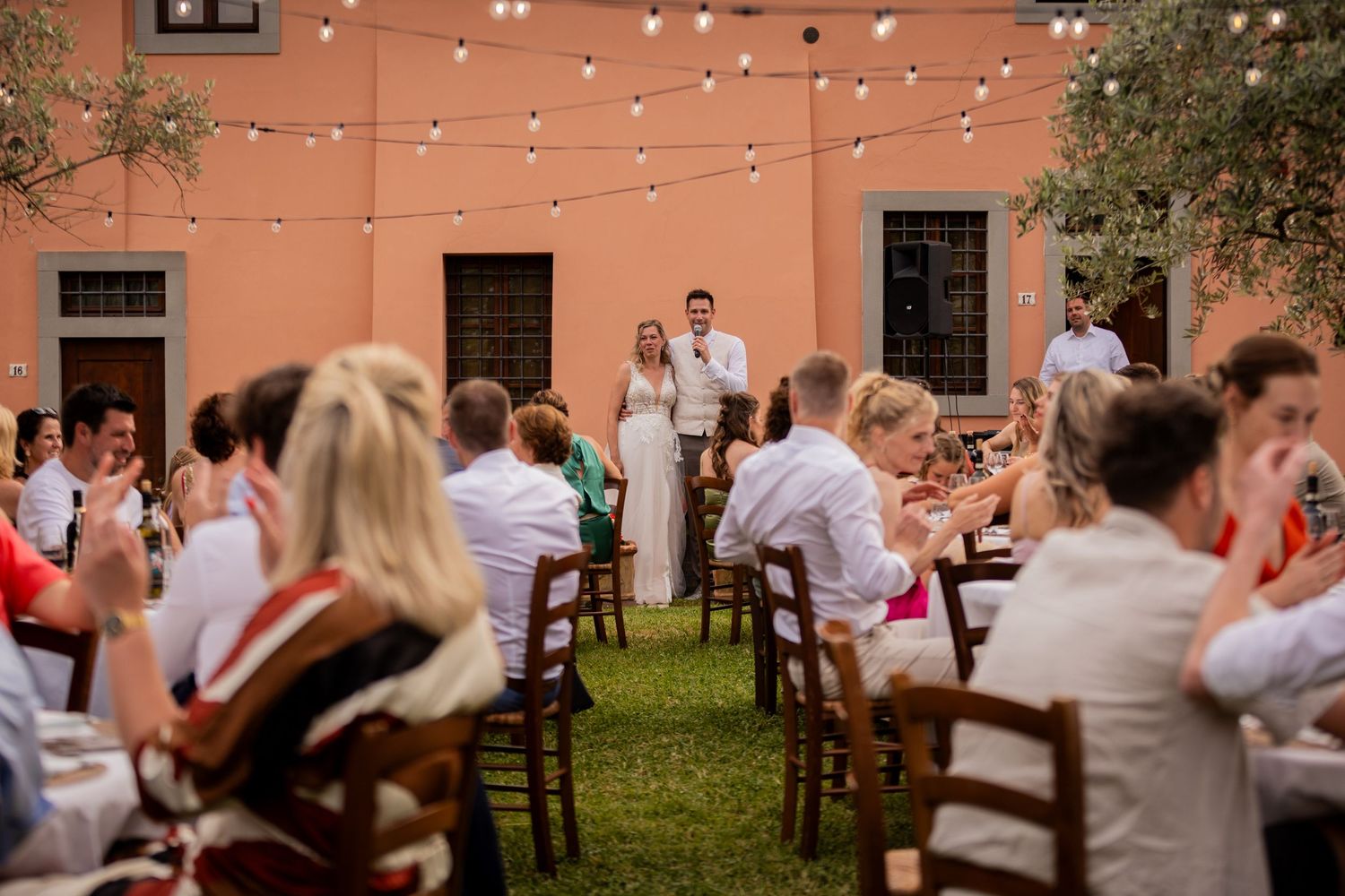 String lights illuminate an outdoor wedding reception with guests seated at tables on a lawn by a terracotta colored building.