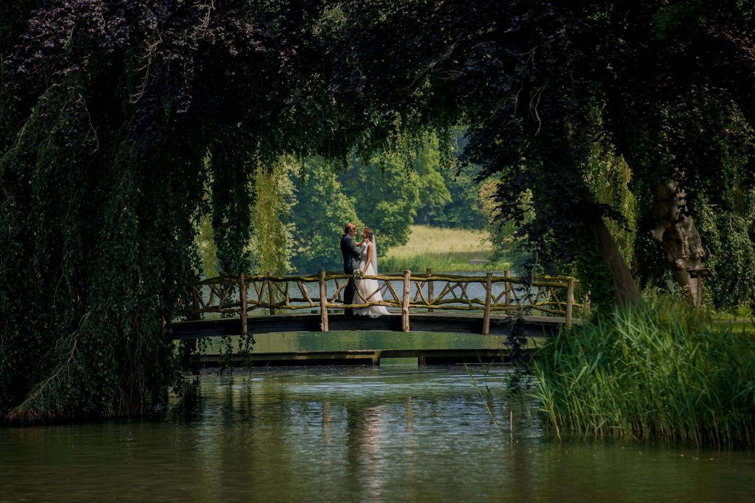 Wedding couple stands romantically on wooden bridge over tranquil pond surrounded by willows.