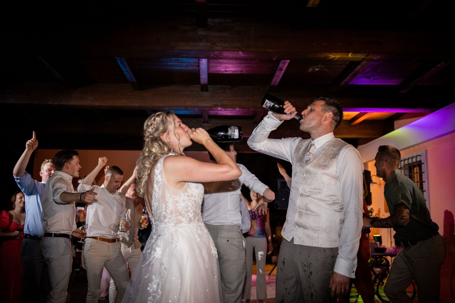 Wedding guests dancing at a reception with colorful lights as people drink and celebrate on the dance floor.